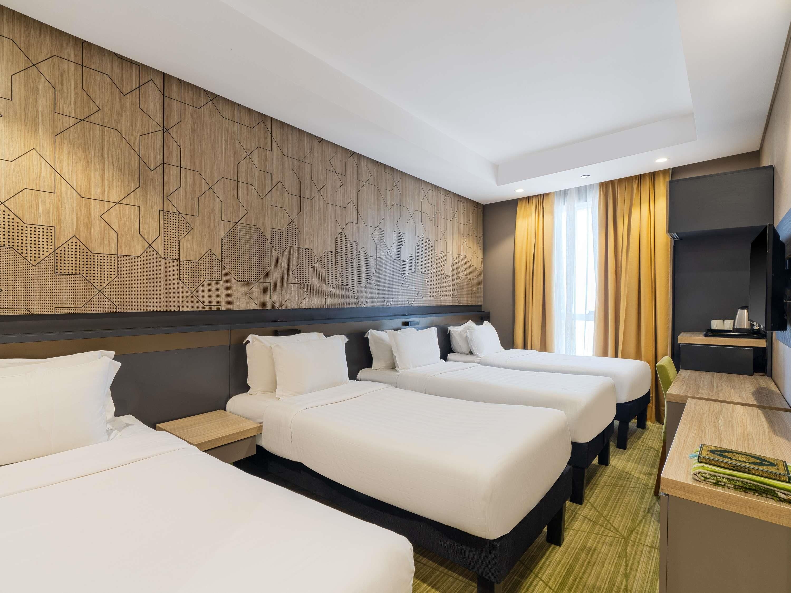 A hotel room with three beds, a wooden accent wall, a television, and a small desk with a lamp.