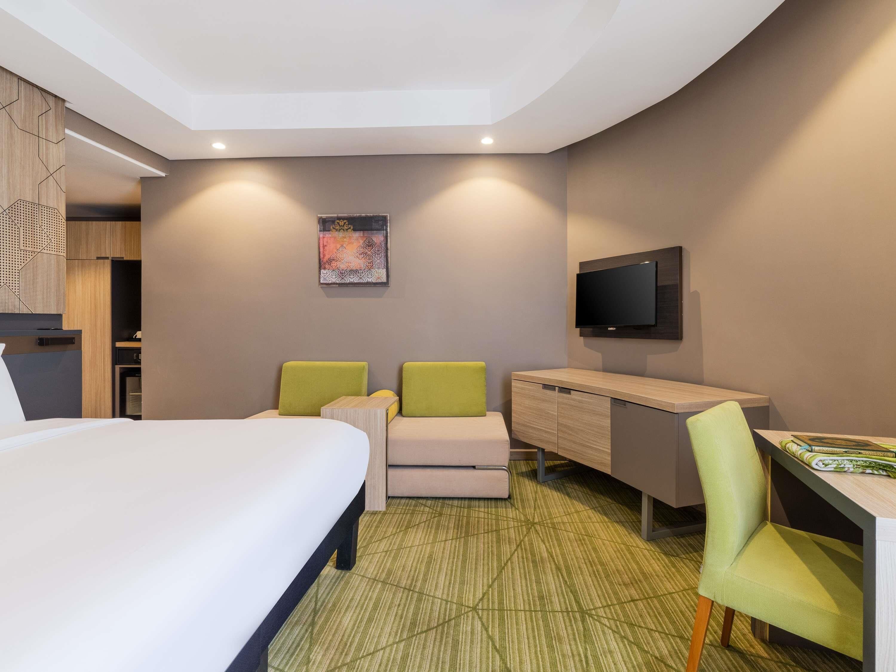 A hotel room with a bed, two green chairs, a desk, a television, and a wall-mounted artwork.