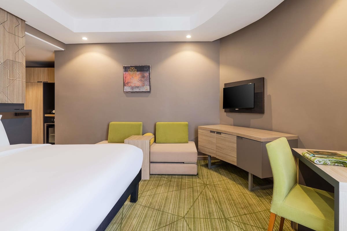 A hotel room with a bed, two green chairs, a desk, a television, and a wall-mounted artwork.
