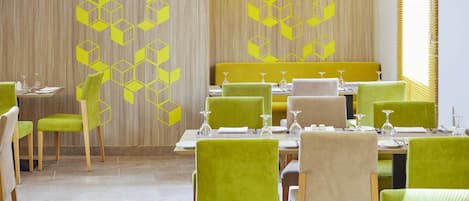 A restaurant with green upholstered chairs, wooden tables, and a wall with a geometric pattern.