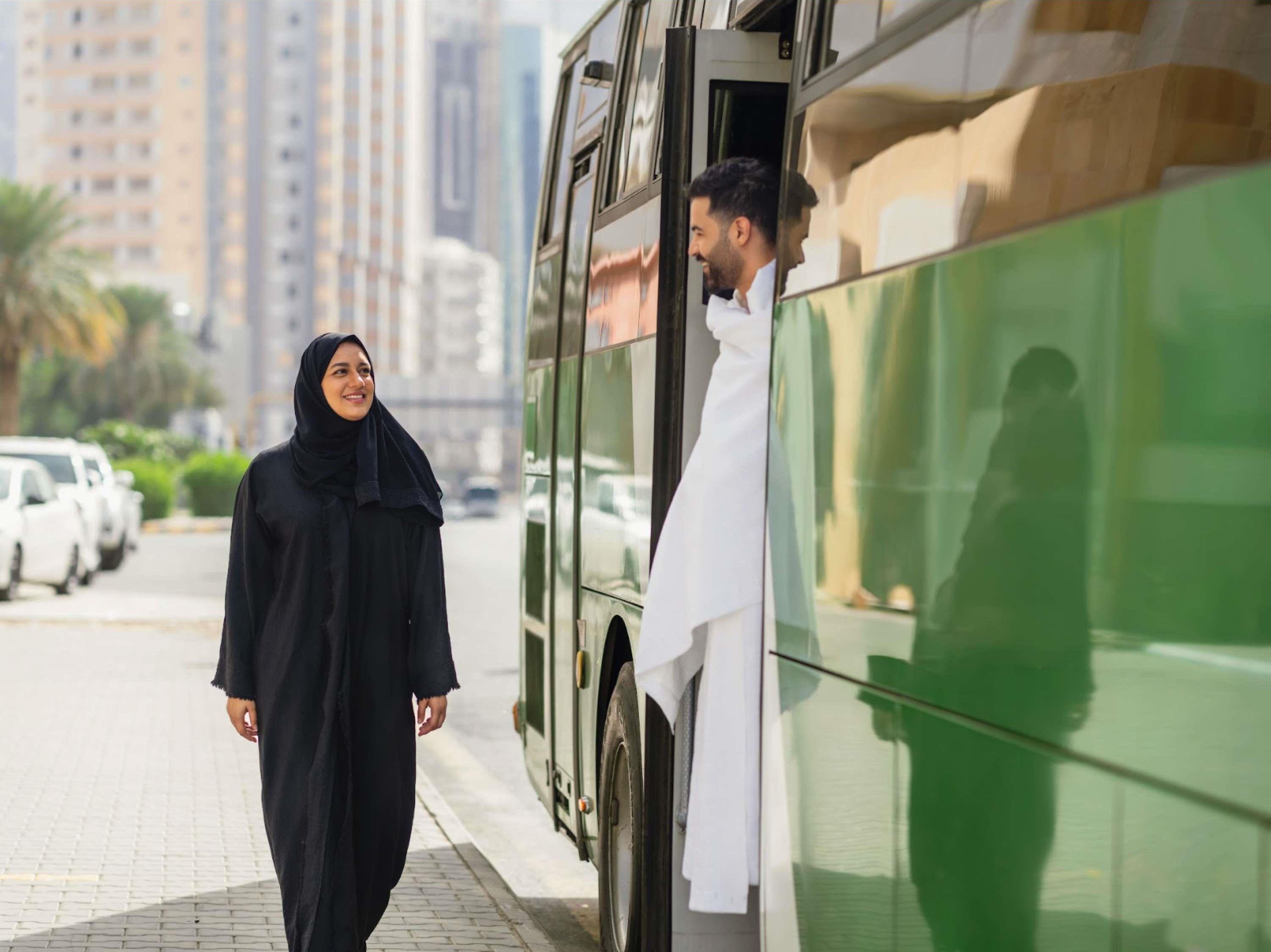 A person in a black dress standing next to a green bus.