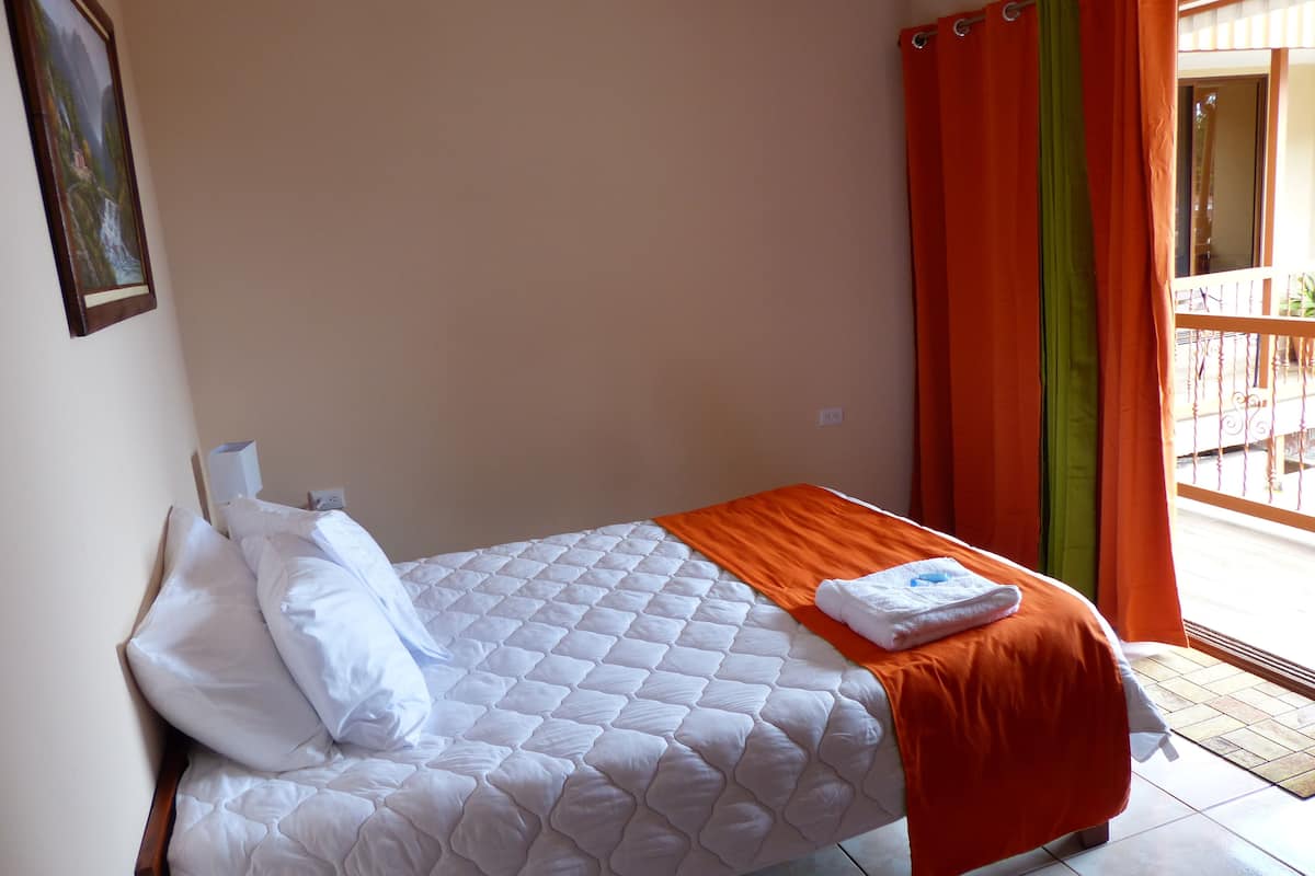 A single bed with white bedding and an orange blanket, a painting on the wall, and orange curtains.