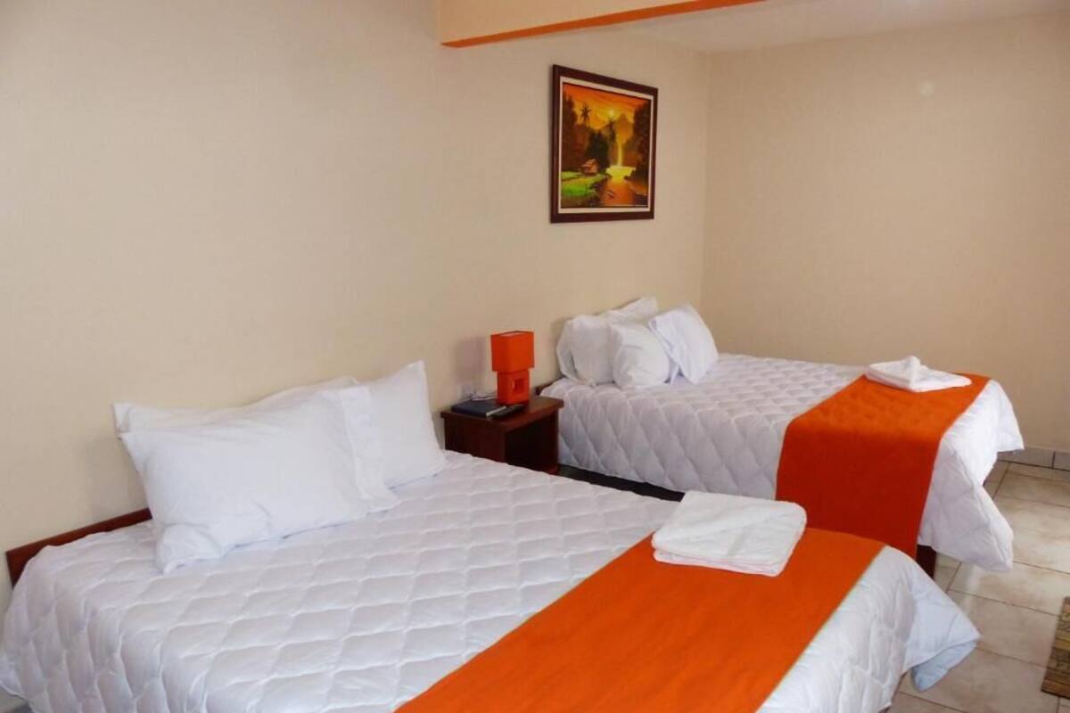 A hotel room with two beds, each with white bedding and orange bedspreads. There is a nightstand with a lamp and a framed picture on the wall.