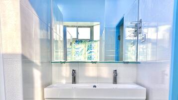 Un baño con un lavamanos blanco, dos llaves y un estante de vidrio sobre el lavamanos.