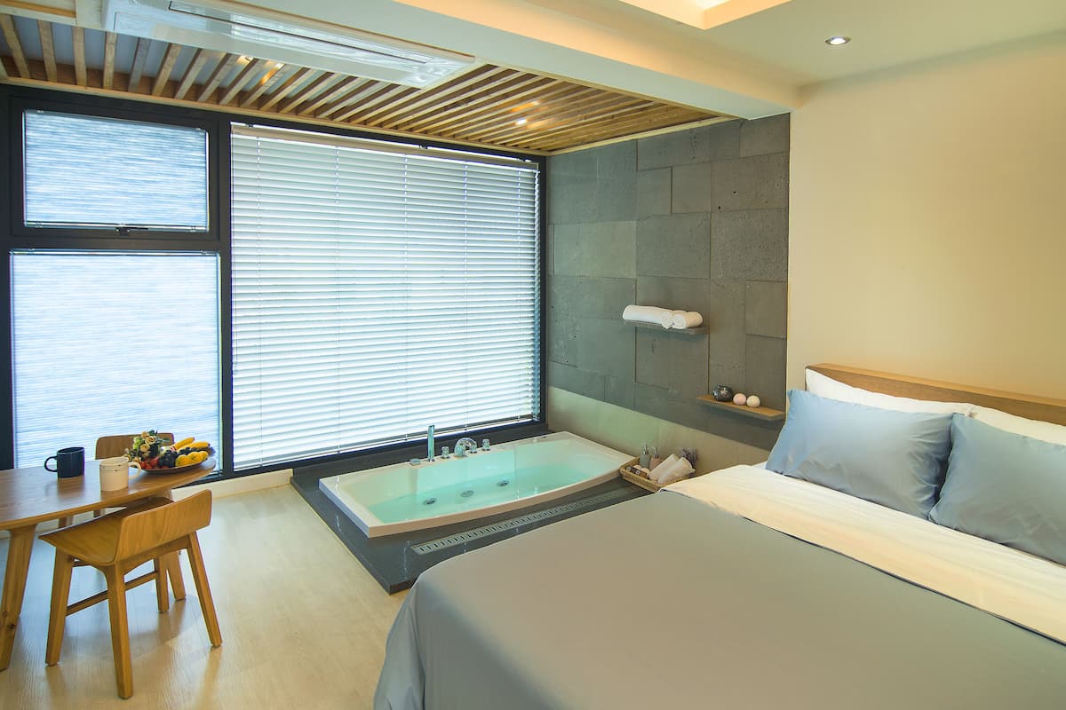 A modern hotel room with a large bed, a bathtub, a dining area with a table and chairs, and a window with blinds.
