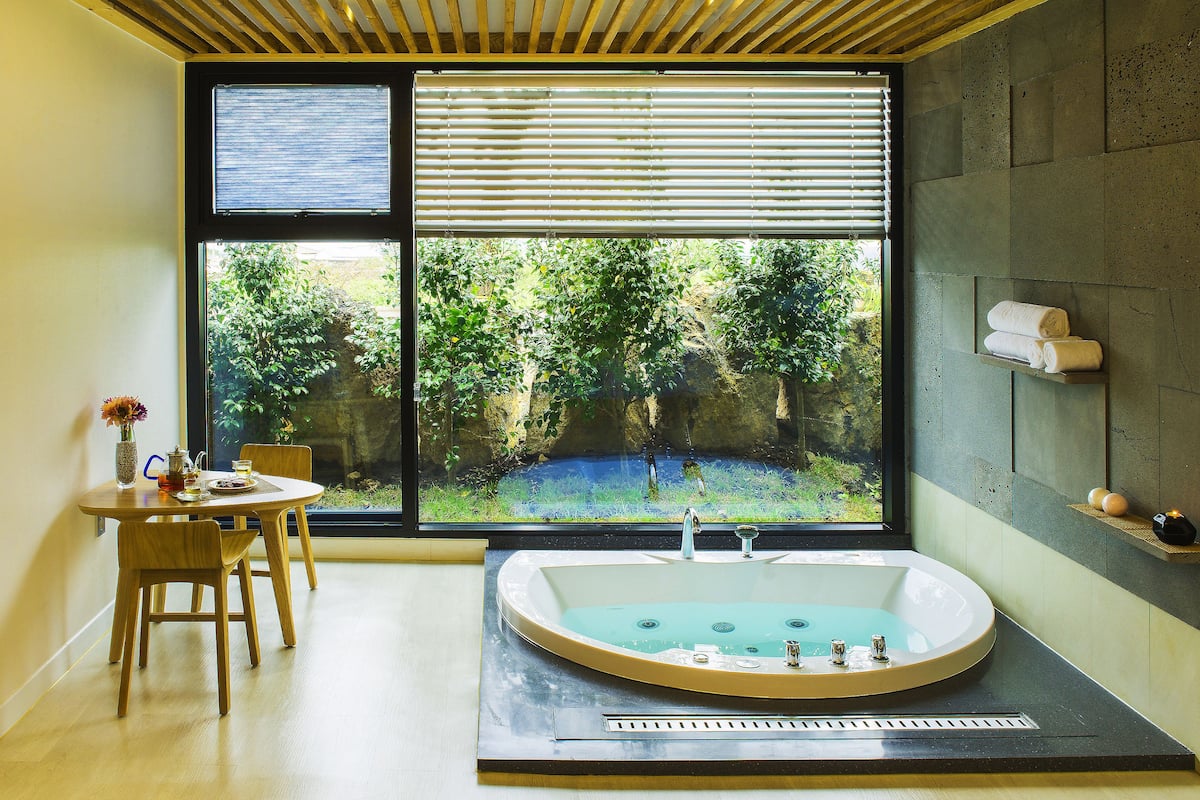 A bathroom with a large window, a bathtub, a sink, and a small table with chairs.