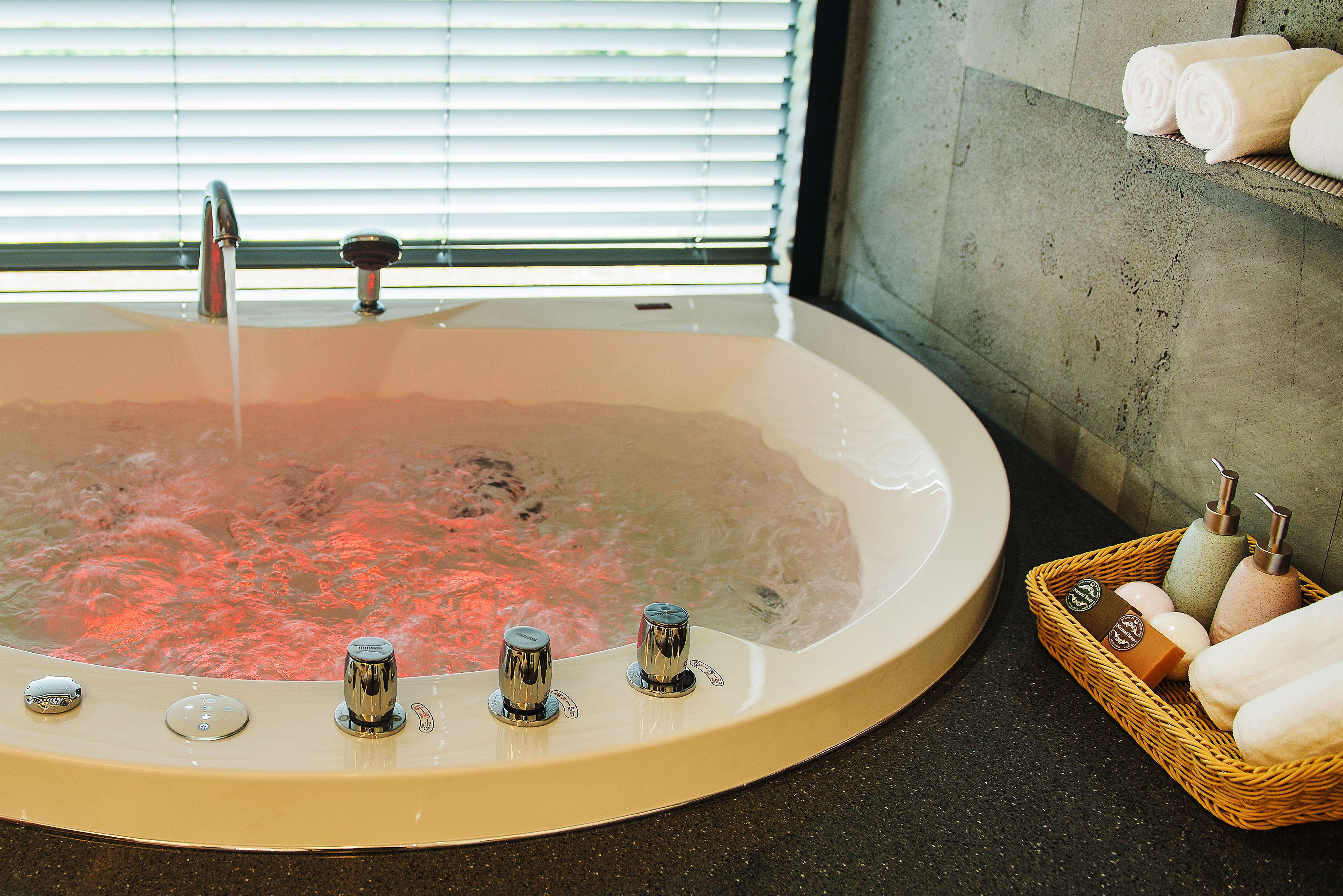 Un jacuzzi con controles, una ventana con persianas y una cesta con toallas y artículos de aseo.