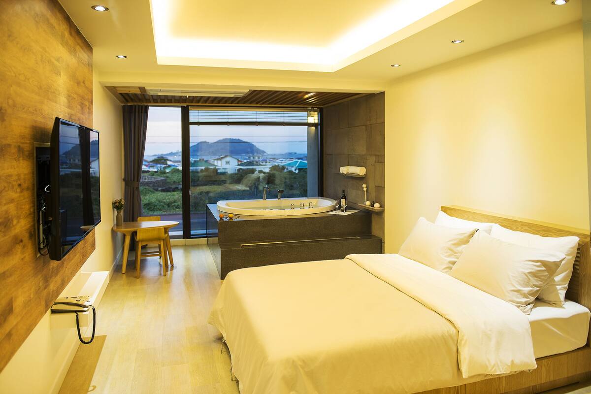 A modern bedroom with a large bed, a television, a desk, and a view of a cityscape through a large window.