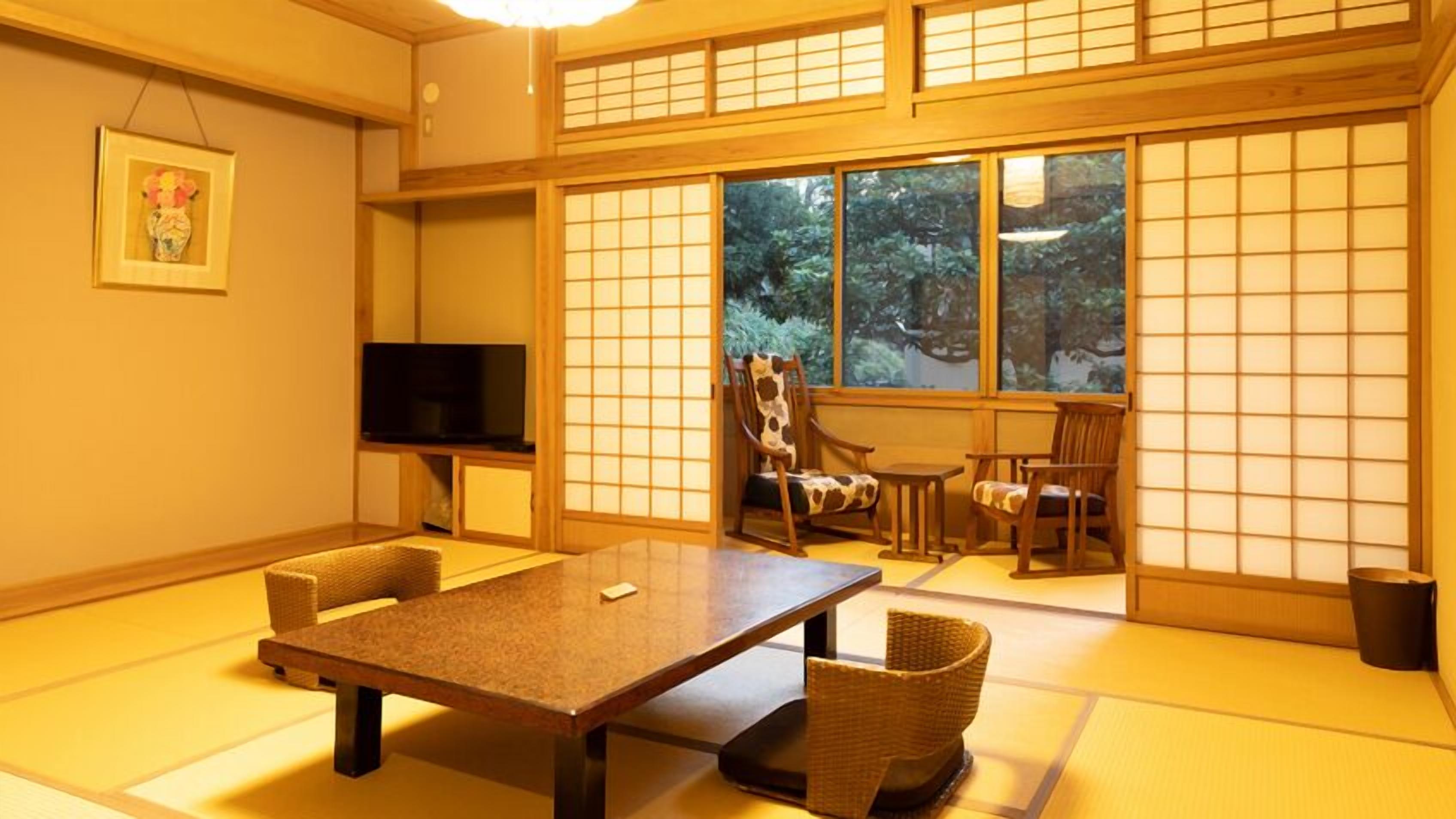 Quarto japonês tradicional com tatames, uma mesa baixa, cadeiras, uma televisão e vista para o verde através de grandes janelas.