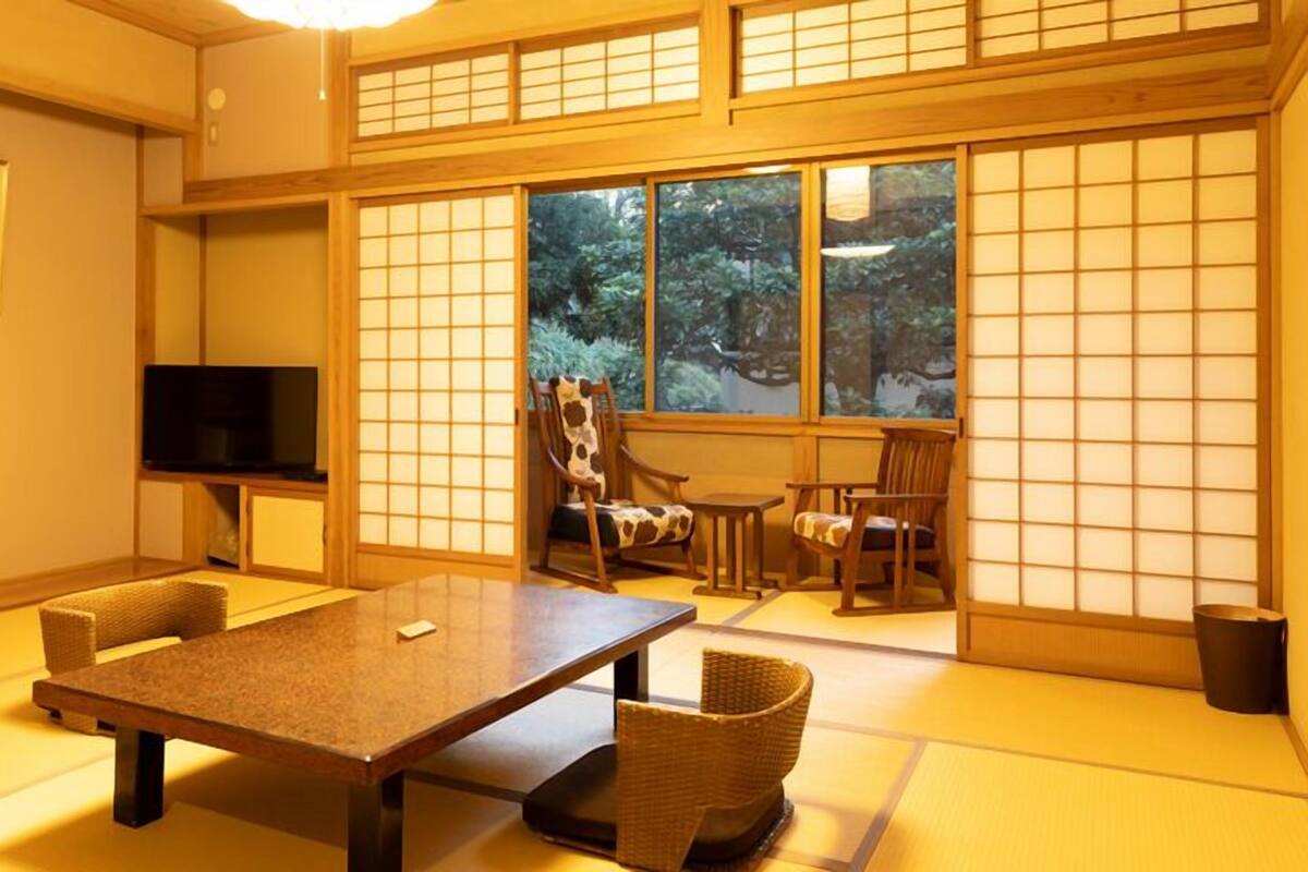 A traditional Japanese room with tatami flooring, a low table, chairs, a television, and a view of greenery through large windows.