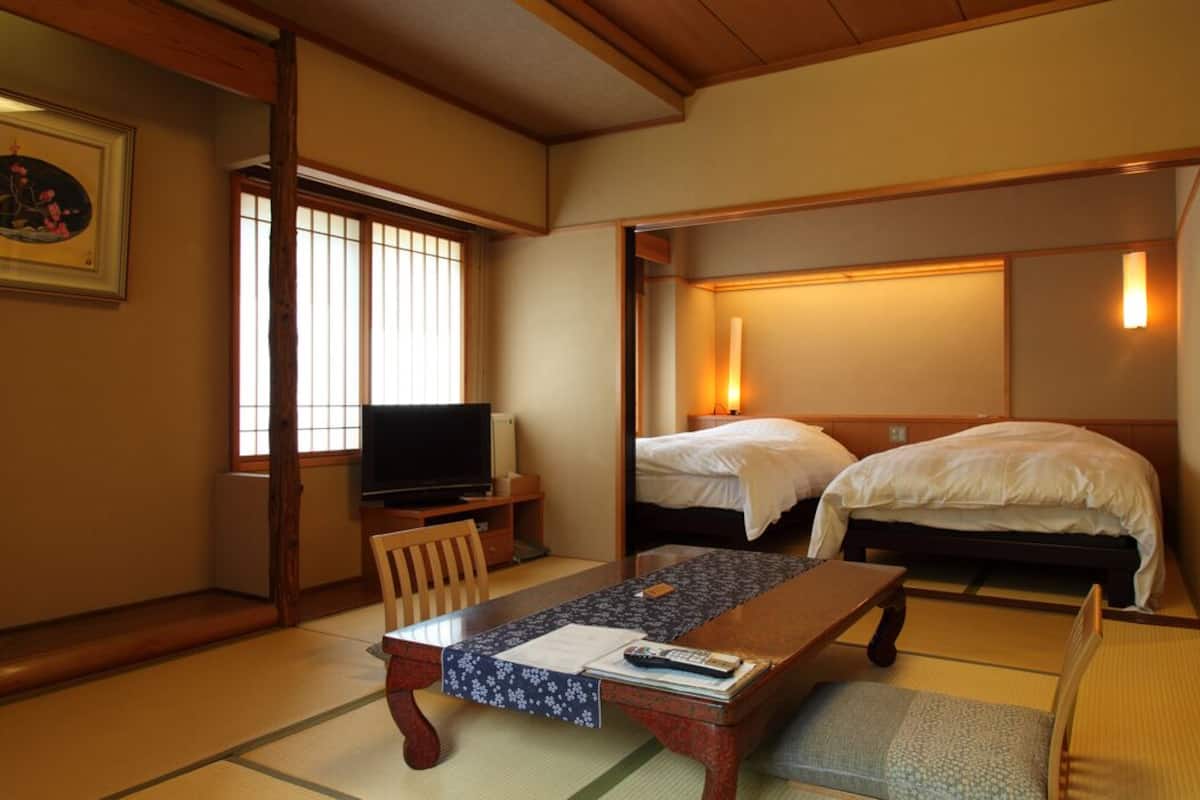 A traditional Japanese room with tatami flooring, a television, two beds, a table with a mat, and a painting on the wall.