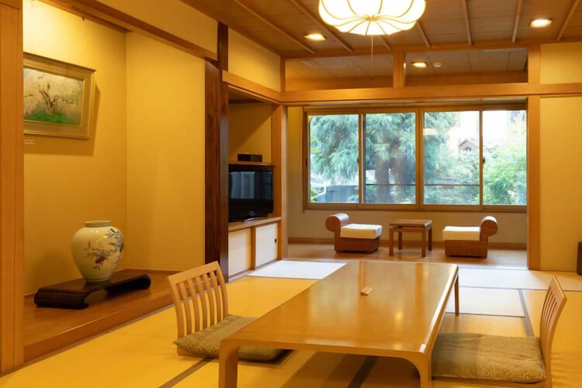 A traditional Japanese-style room with tatami flooring, a low table, cushions, a television, and a large window with a view of trees.
