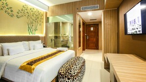 Quarto de hotel com uma cama grande, um banco de madeira, uma televisão e um box de chuveiro em vidro.