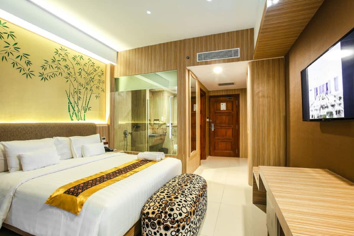 A hotel room with a large bed, a wooden bench, a television, and a glass shower enclosure.