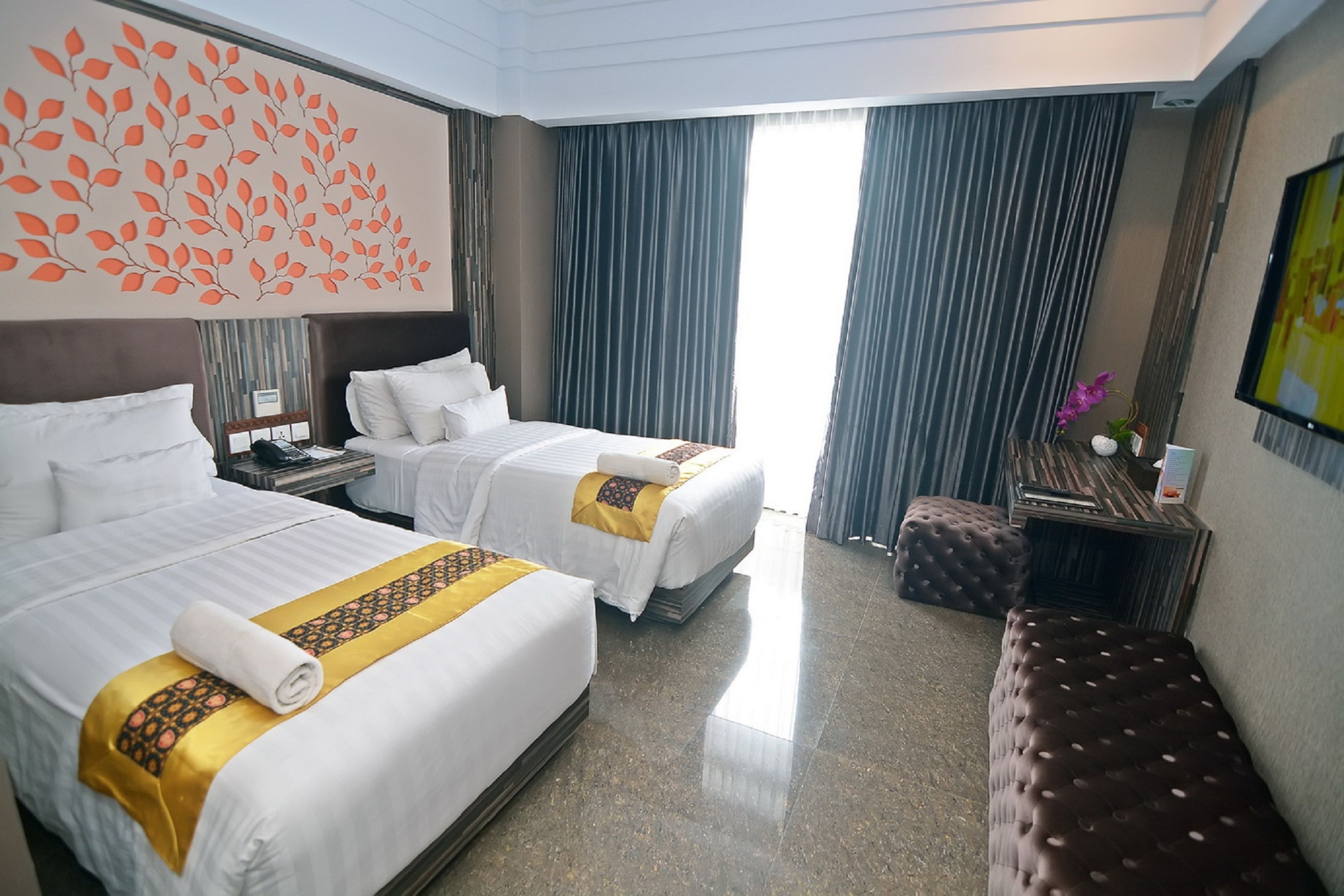 Ein Hotelzimmer mit zwei Betten, einer Bank, einem Schreibtisch und einem großen Fenster mit Vorhängen.
