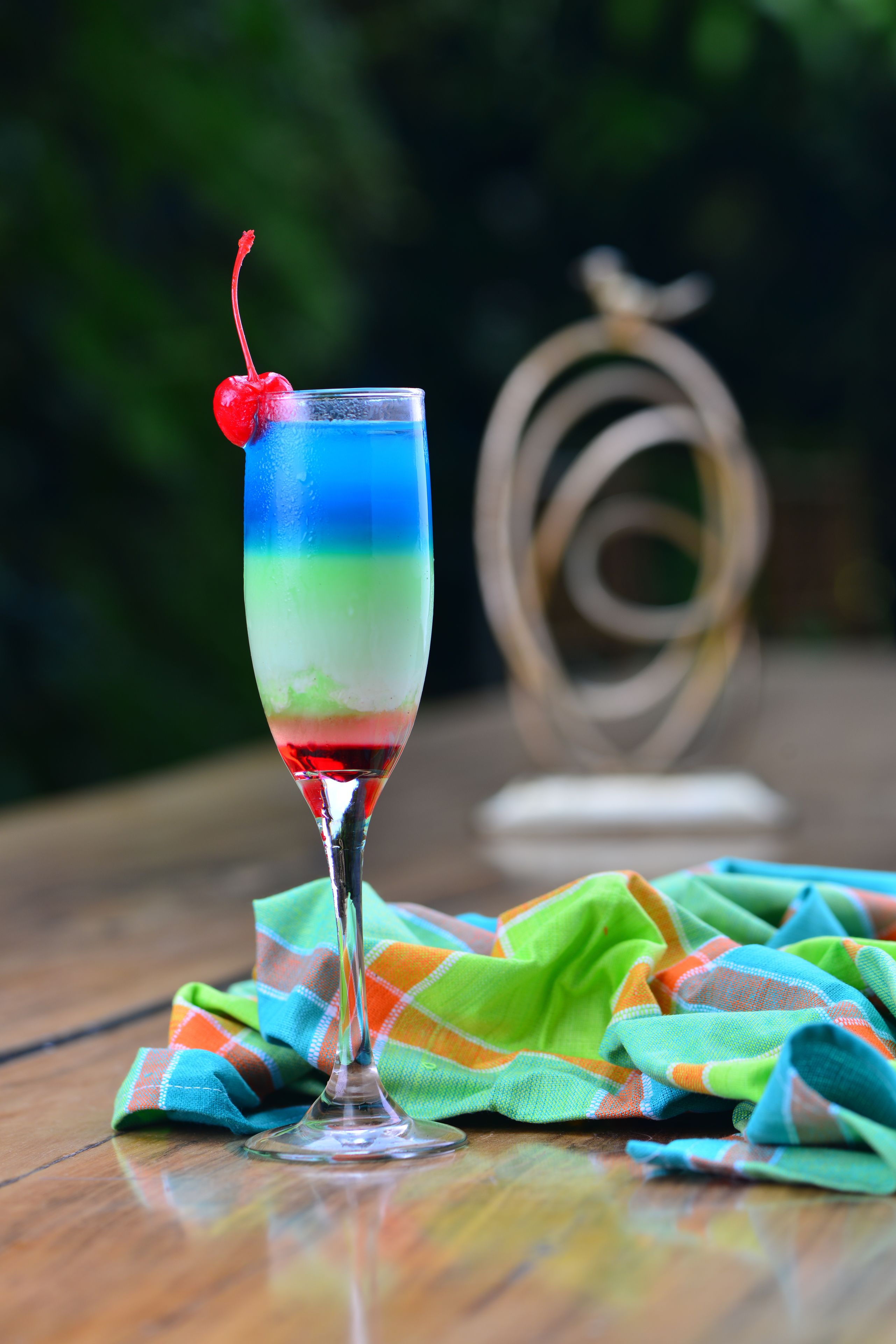 A glass with a layered drink and a cherry on top, placed on a wooden table with a colorful cloth.