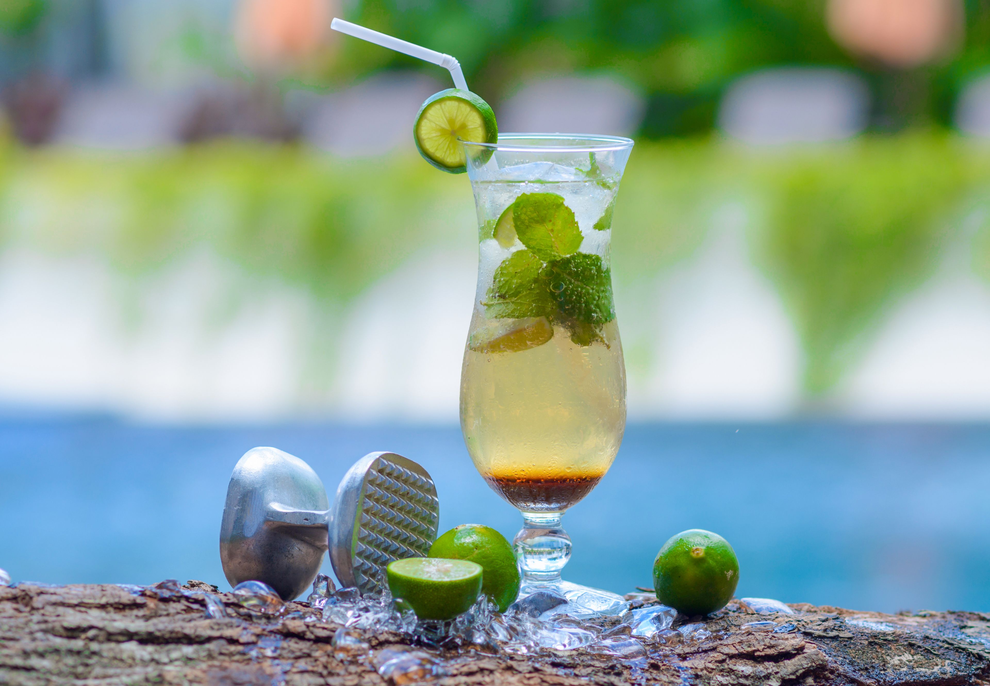 A glass of drink with lime and mint garnish, ice cubes, and a lime wedge on a wooden surface with a blurred outdoor background.