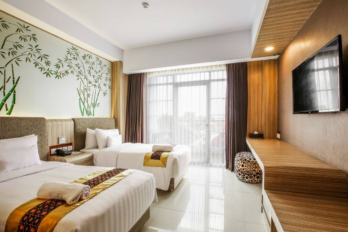 A hotel room with two beds, a large window with curtains, a television on the wall, and a wooden bench.
