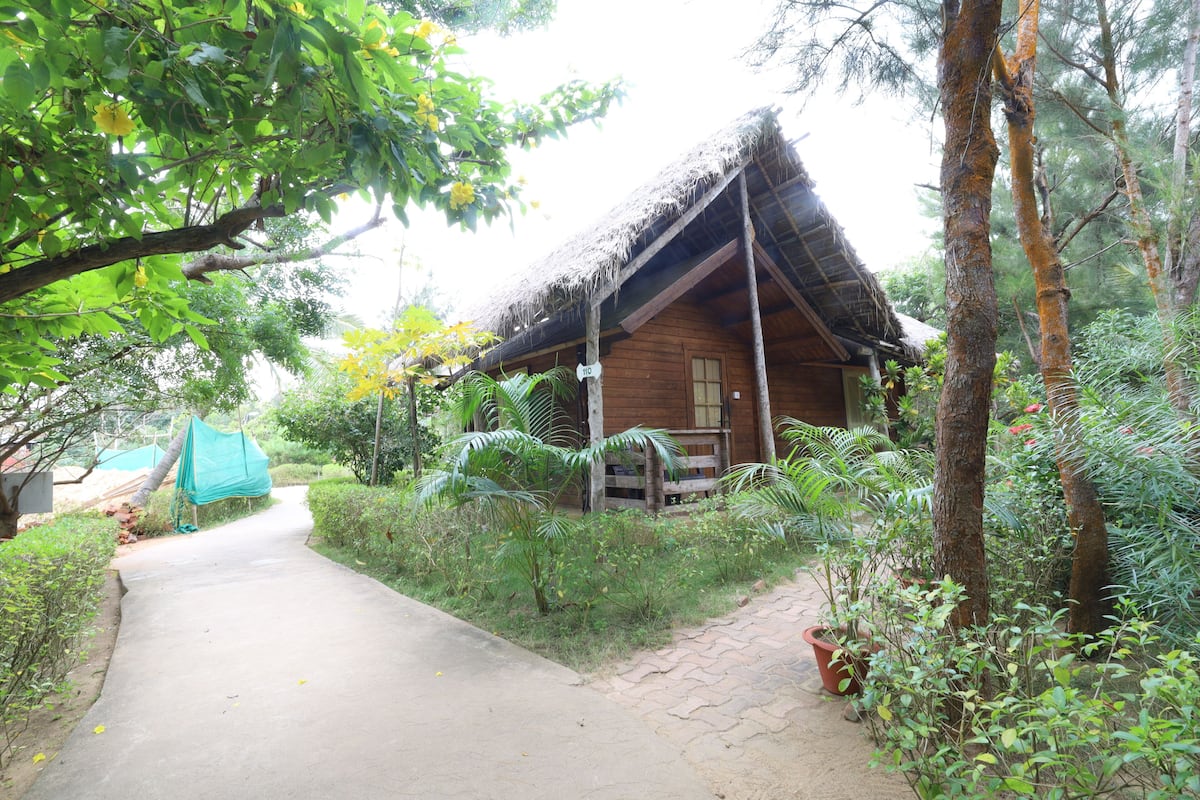 A wooden cabin with a thatched roof, surrounded by lush greenery and a paved path leading to it.