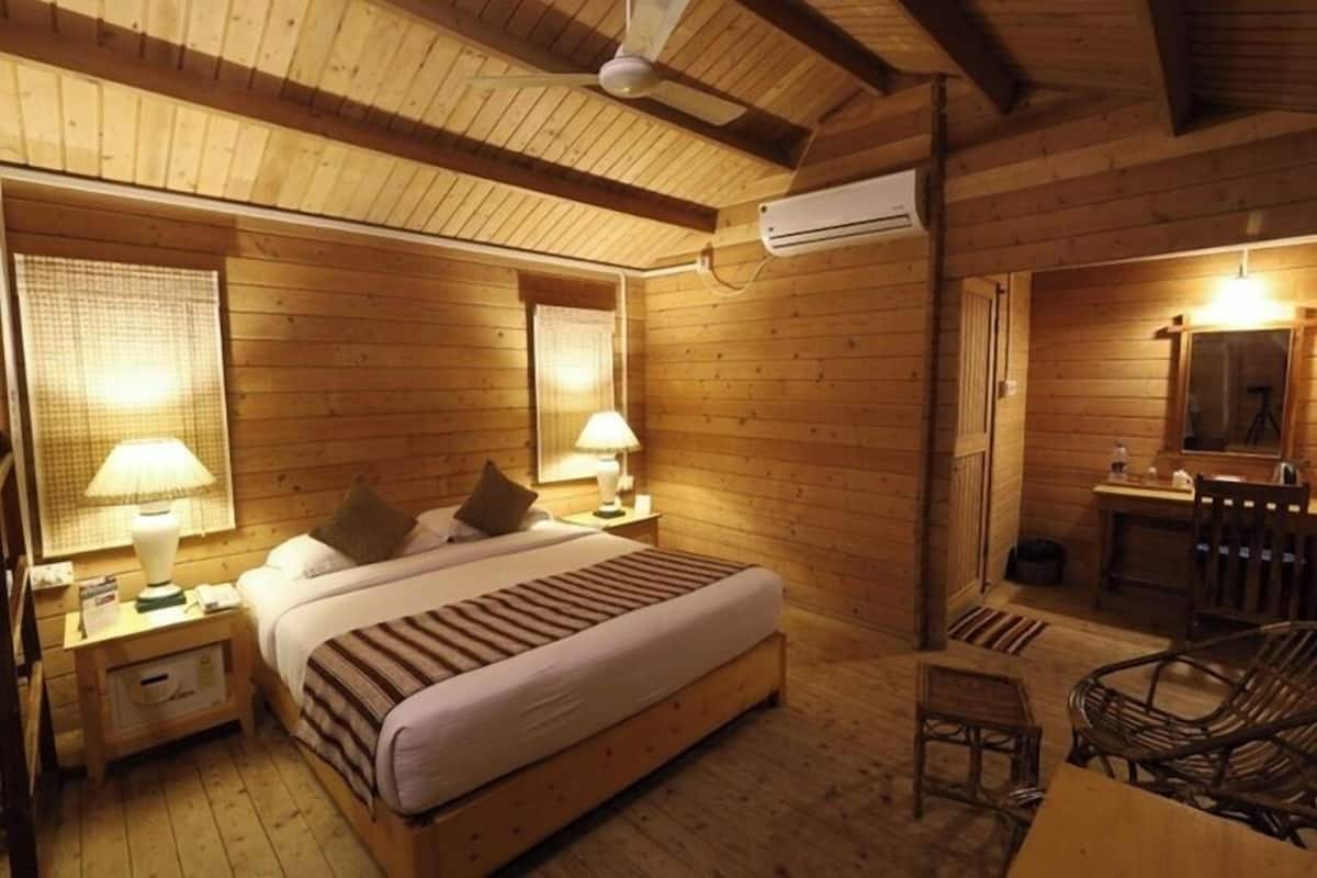 A wooden cabin room with a bed, two bedside lamps, a chair, a small table, and a mirror.