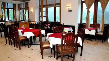 A restaurant with multiple tables, each covered with a white tablecloth and red napkins. The room has large windows with curtains, and the floor has a herringbone pattern.