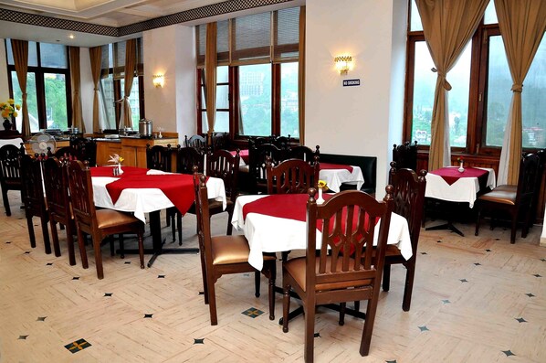 A restaurant with multiple tables, each covered with a white tablecloth and red napkins. The room has large windows with curtains, and the floor has a herringbone pattern. - Hotel Asia The Dawn (Shimla)