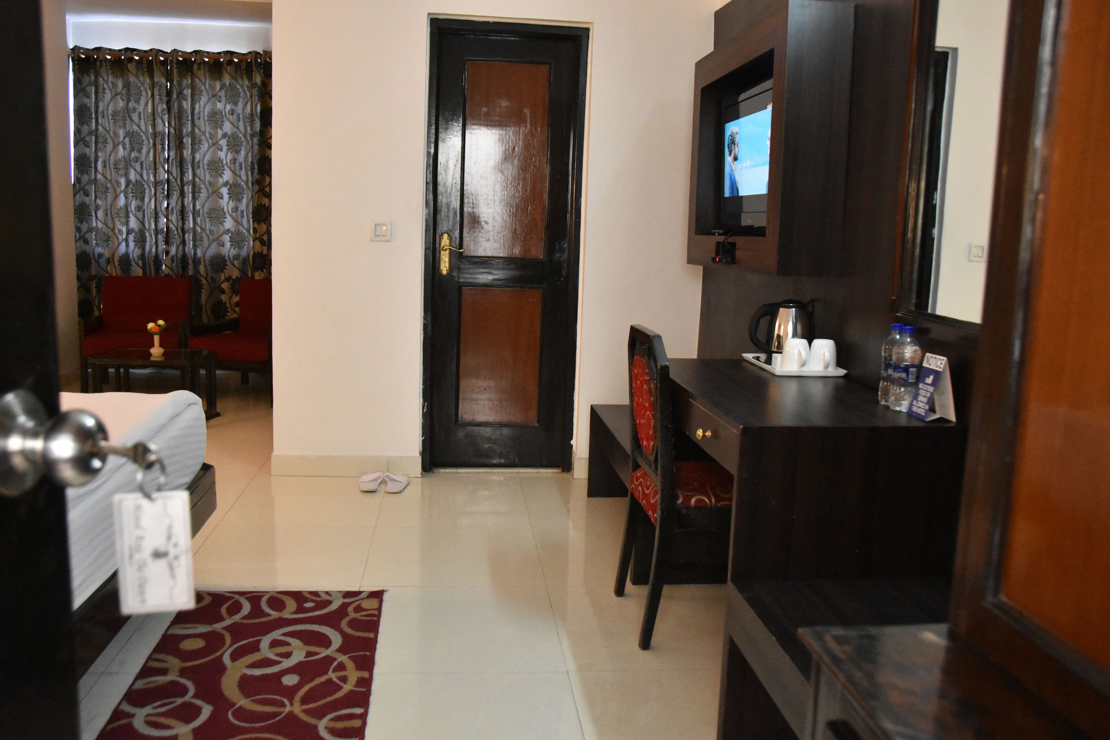 A hotel room with a wooden door, a television mounted on the wall, a small table with a vase, and a red patterned rug.