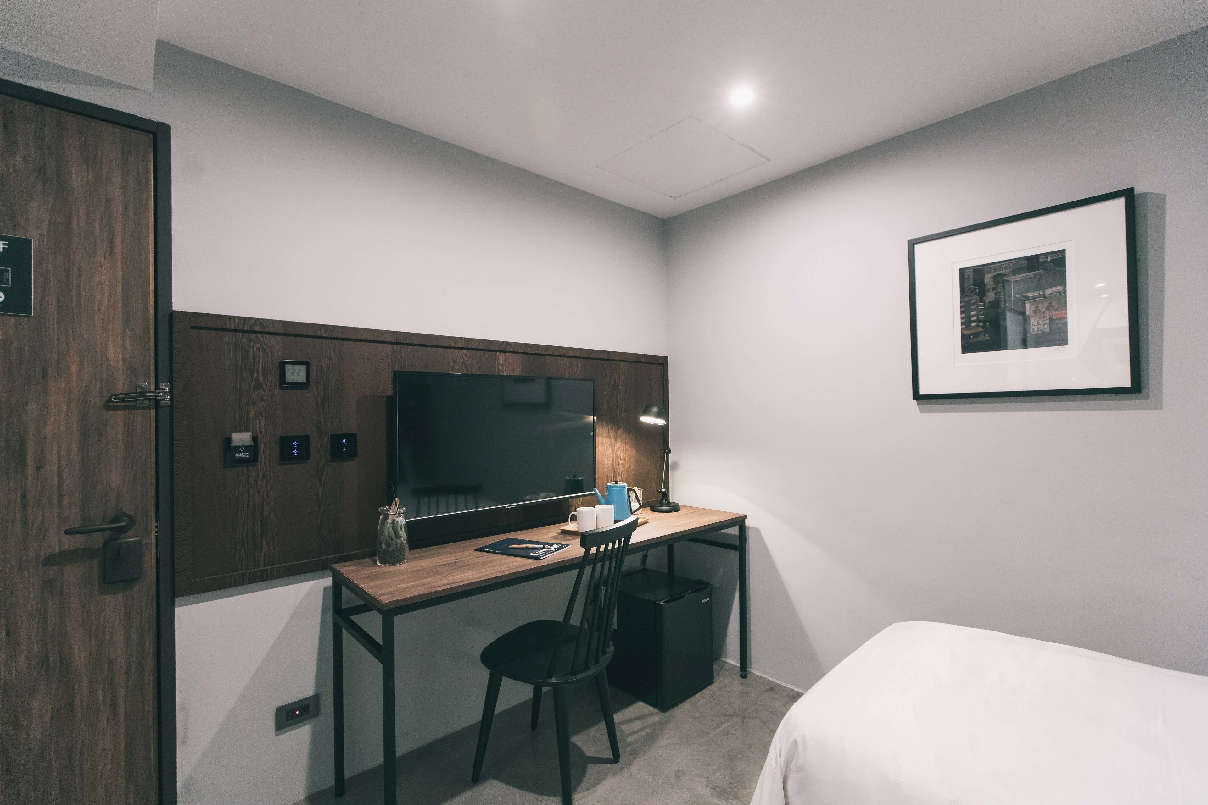 A compact hotel room with a wooden desk, a chair, a bed, a small trash bin, and a framed picture on the wall.