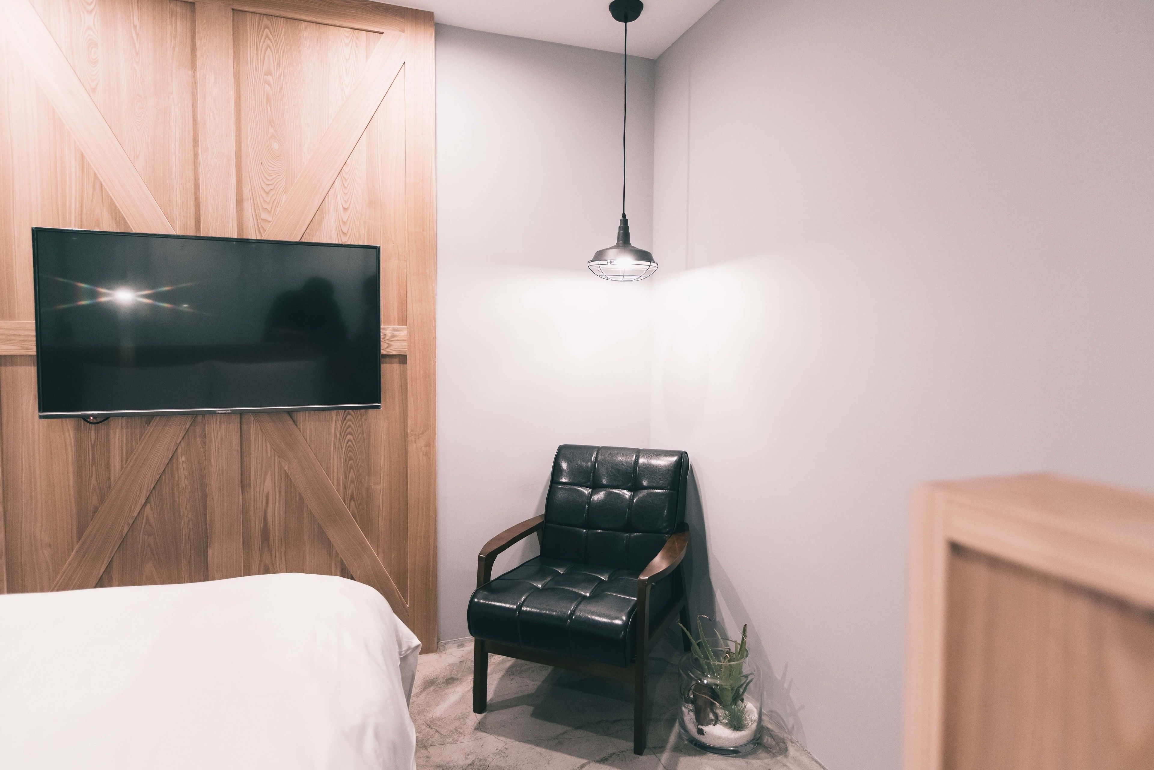 Ein Schlafzimmer mit einem Bett, einem schwarzen Ledersessel, einem an einer Holzwand montierten Fernseher und einer Pendelleuchte.