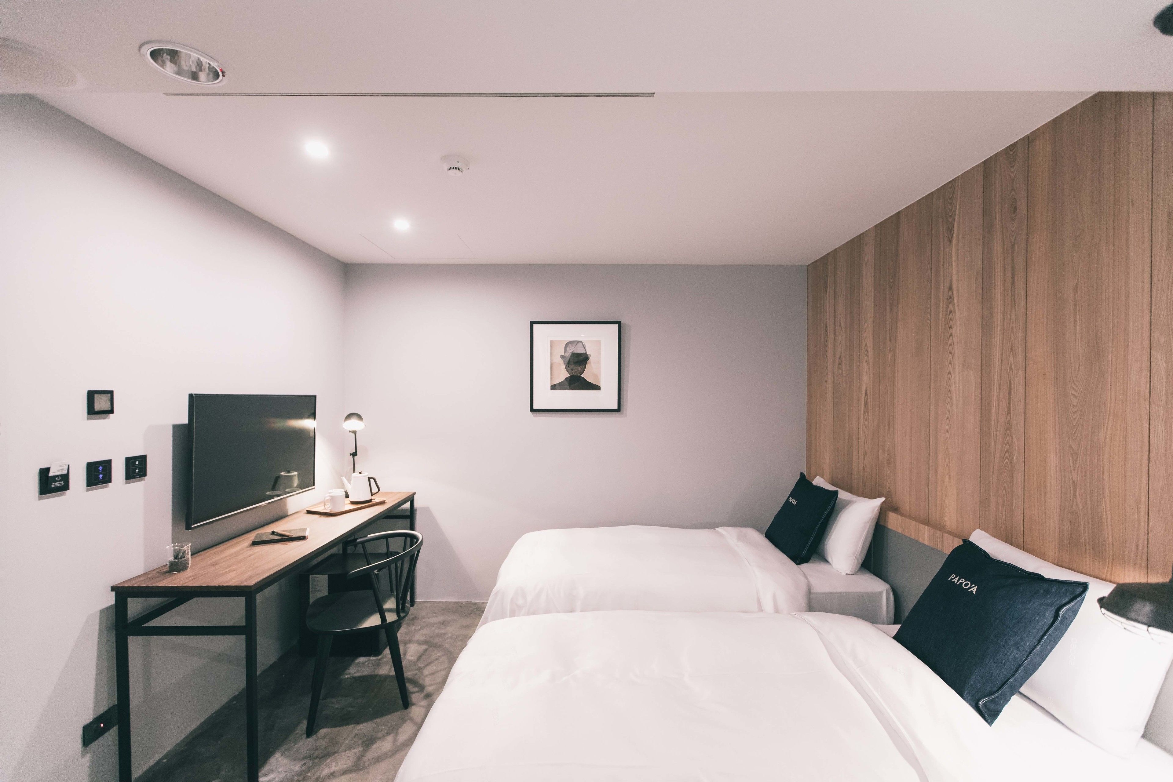 A modern hotel room with two beds, a desk with a computer, a chair, and a wooden accent wall.