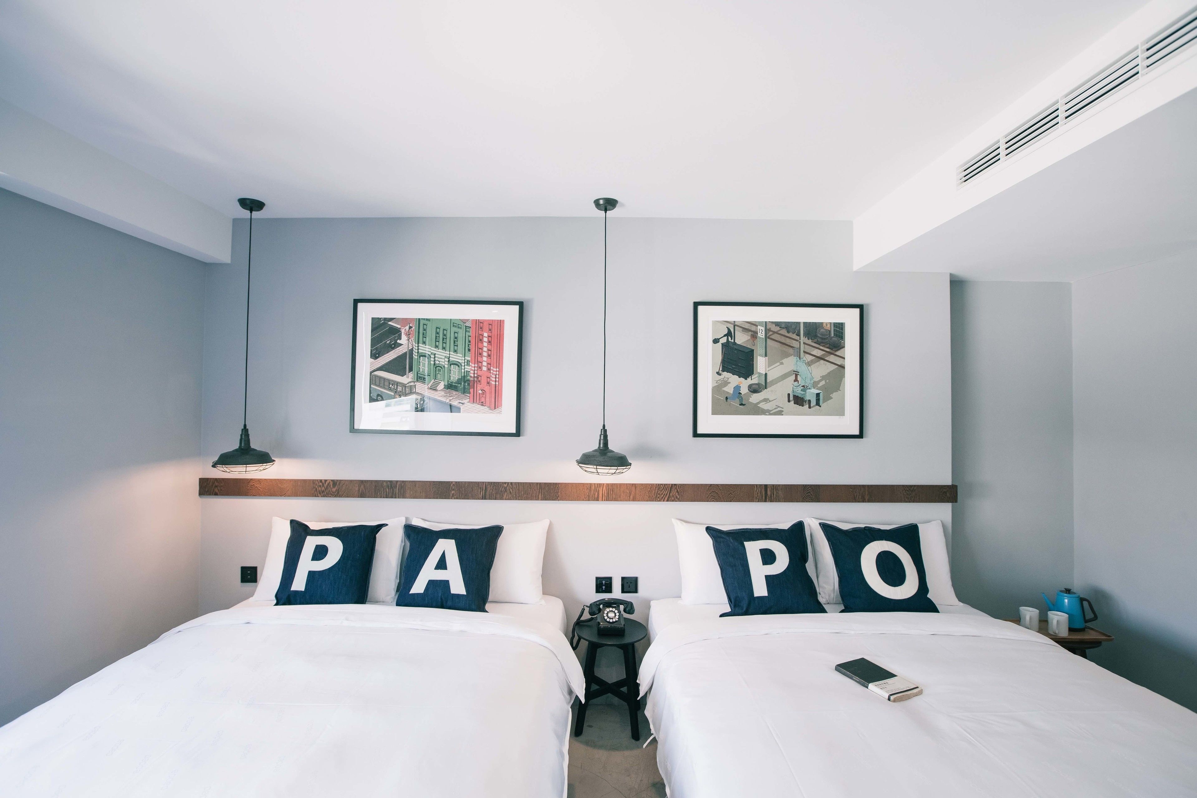 A hotel room with two beds, each with a pillow featuring a large letter. There are framed pictures on the wall, a small table with a telephone, and a kettle on a side table.