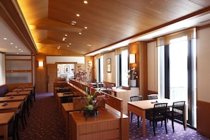 A hotel dining area with wooden tables and chairs, a long counter, and large windows.