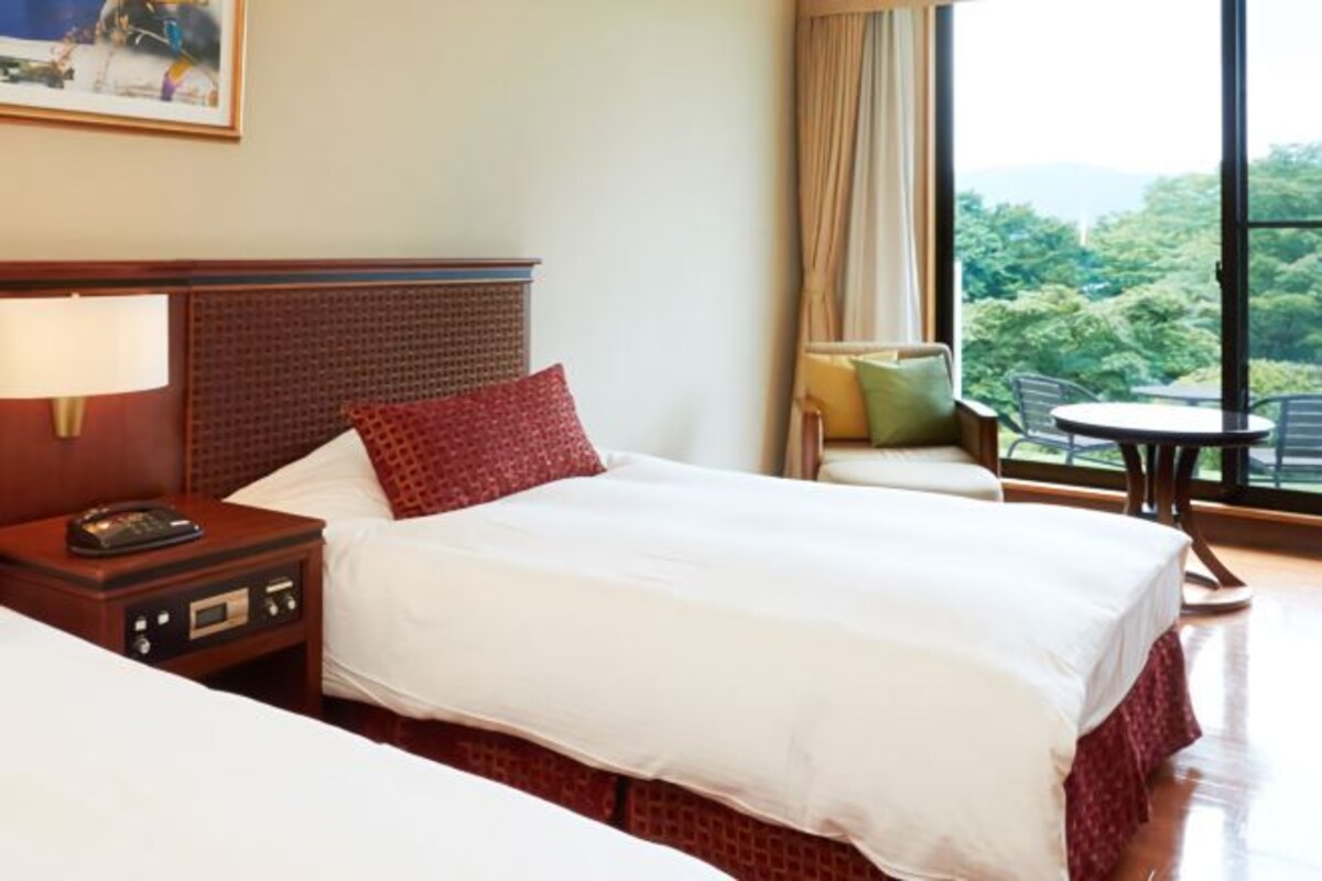 Habitación de hotel con una cama grande, una mesita de noche con lámpara, una silla y vistas al exterior.