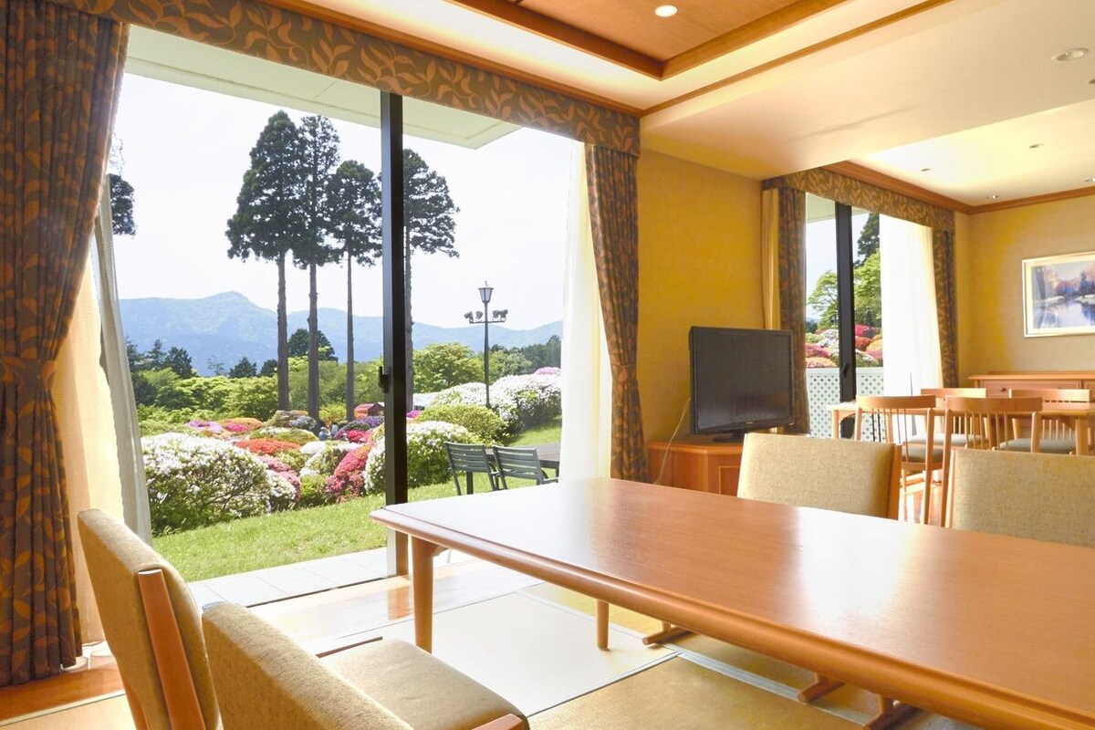 Una camera d'albergo con zona pranzo, televisione e vista su un giardino con alberi e fiori.