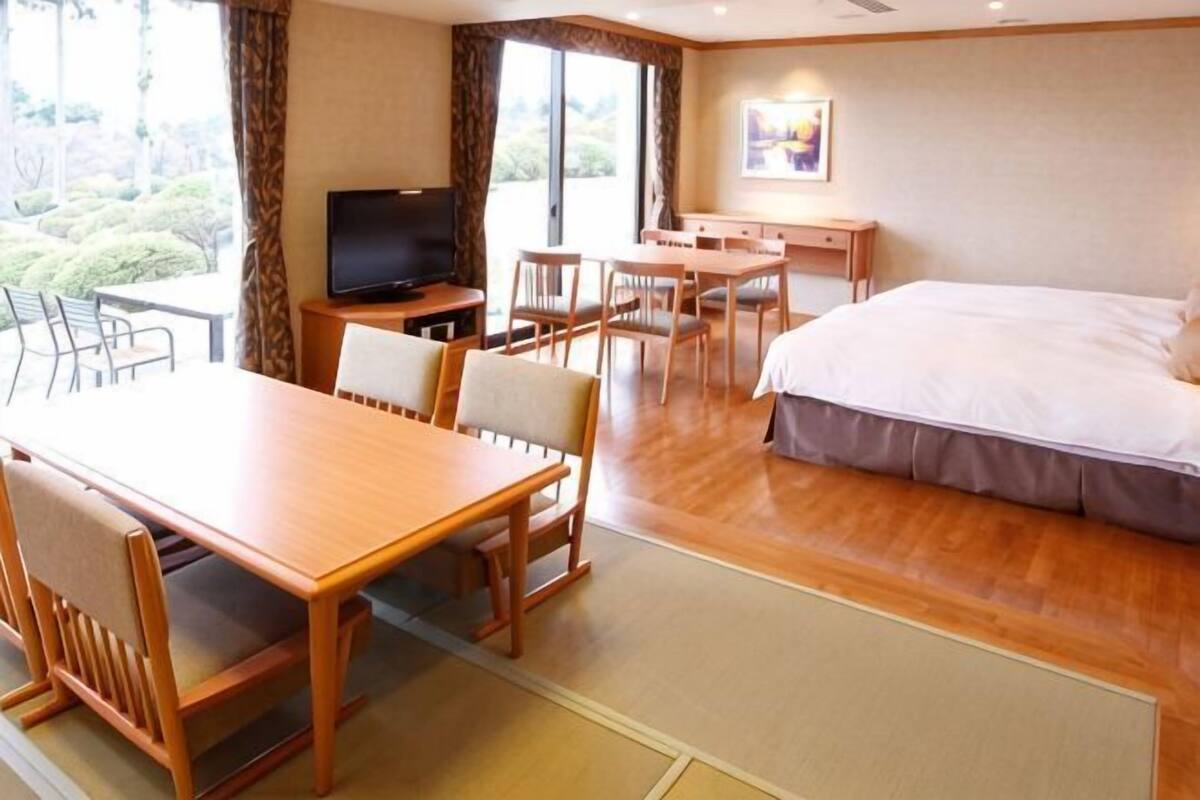 Habitación de hotel con cama, televisión, mesa de comedor, sillas y vista al exterior.