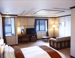 Habitación de hotel con cama, escritorio con televisor, silla, mesita y ventana.