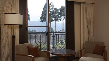 A hotel room with two chairs, a small round table, a large window with a view of trees and hills, and a floor lamp.