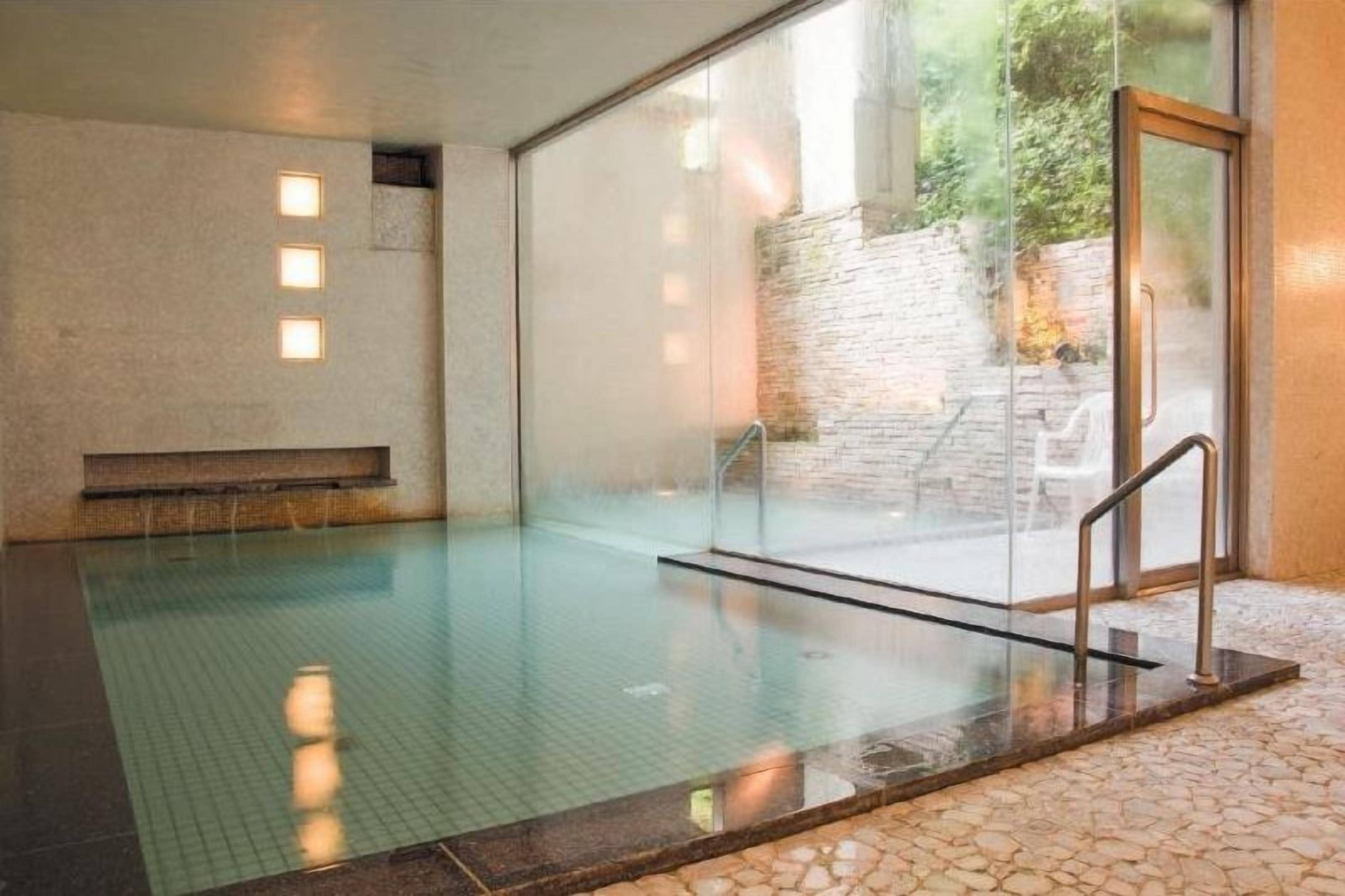 A clear indoor swimming pool with a glass wall, a stone wall outside, and a metal handrail.