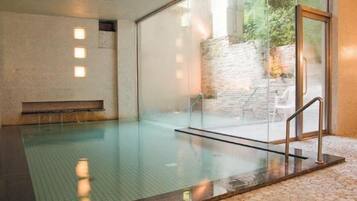 A clear indoor swimming pool with a glass wall, a stone wall outside, and a metal handrail.