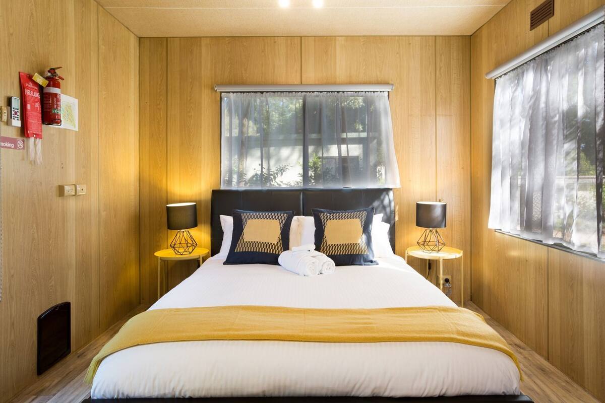 A bedroom with a wooden paneled wall, a bed with a white and yellow bedspread, two bedside tables with lamps, a window with sheer curtains, and a fire extinguisher on the wall.