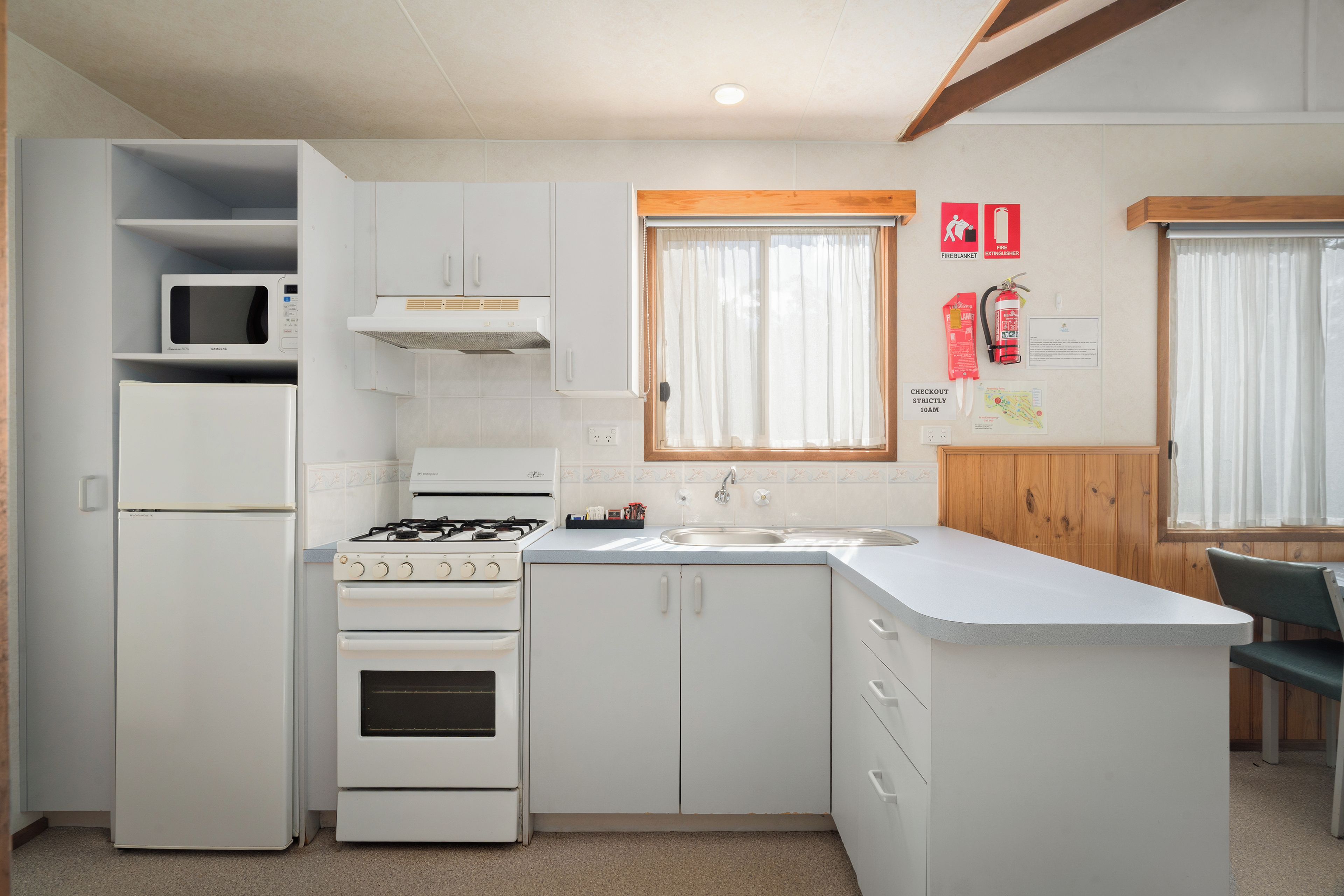 A compact kitchen with white appliances, a built-in microwave, a gas stove, and a sink. There are cabinets and a window with curtains.