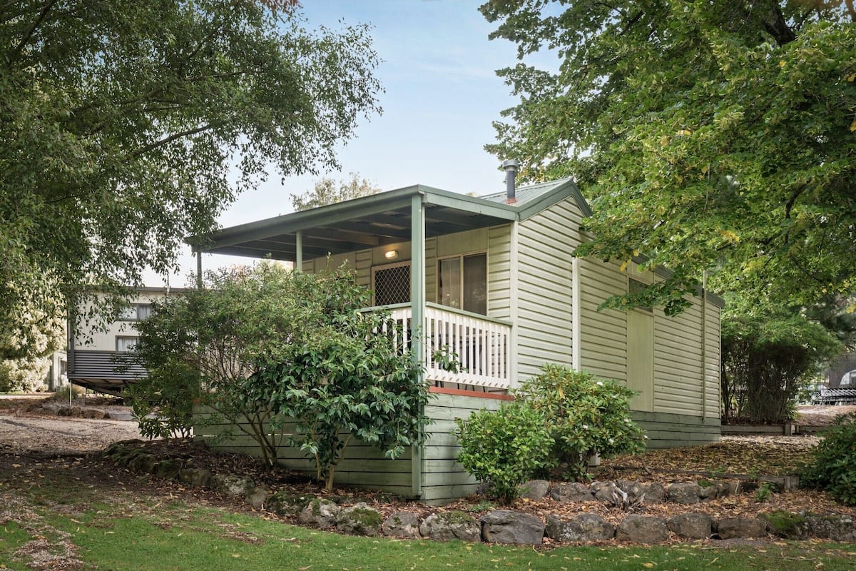 A small, single-story house with a porch, surrounded by trees and a grassy yard.