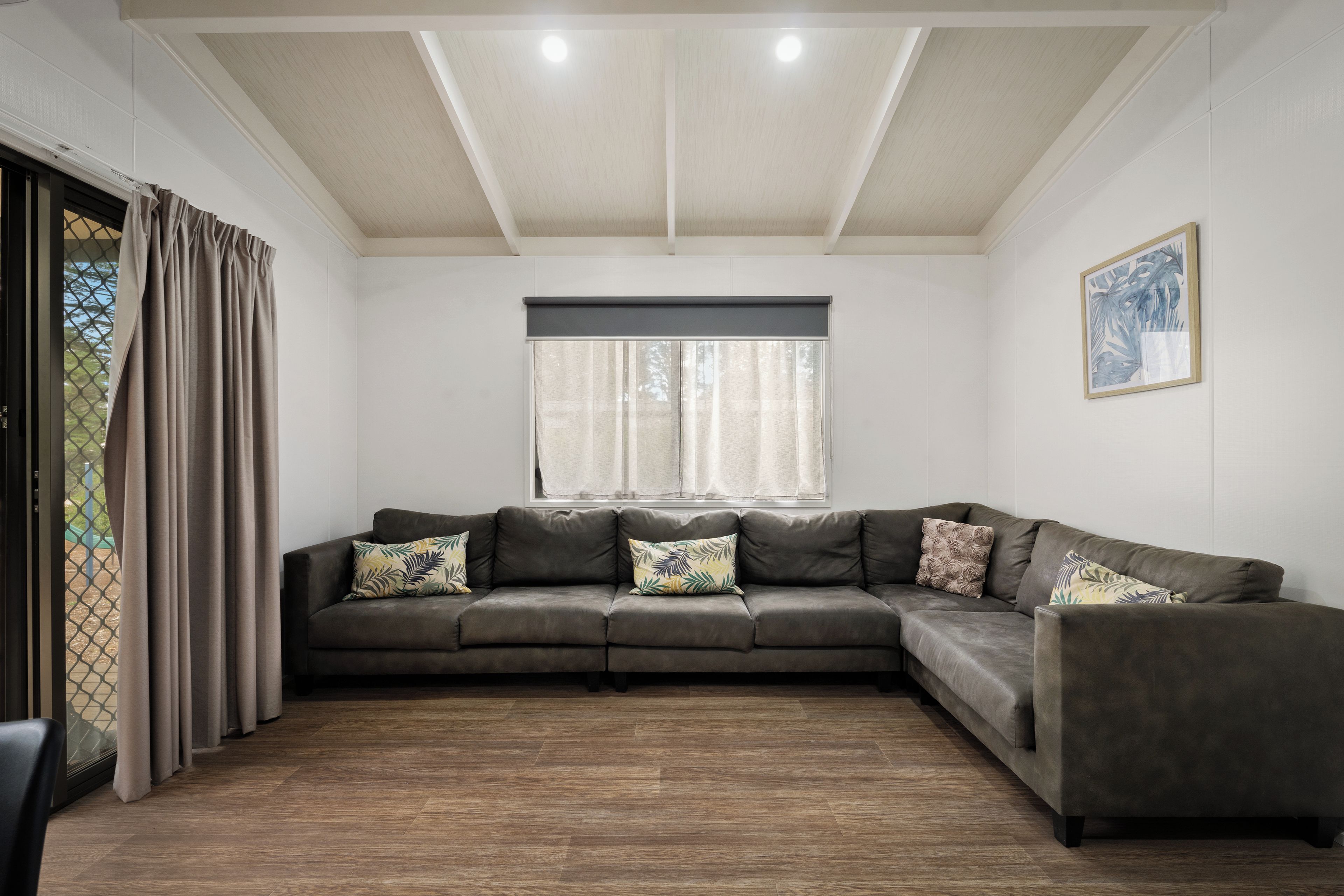 A modern living room with a sectional sofa, a window with curtains, a framed picture on the wall, and a wooden floor.