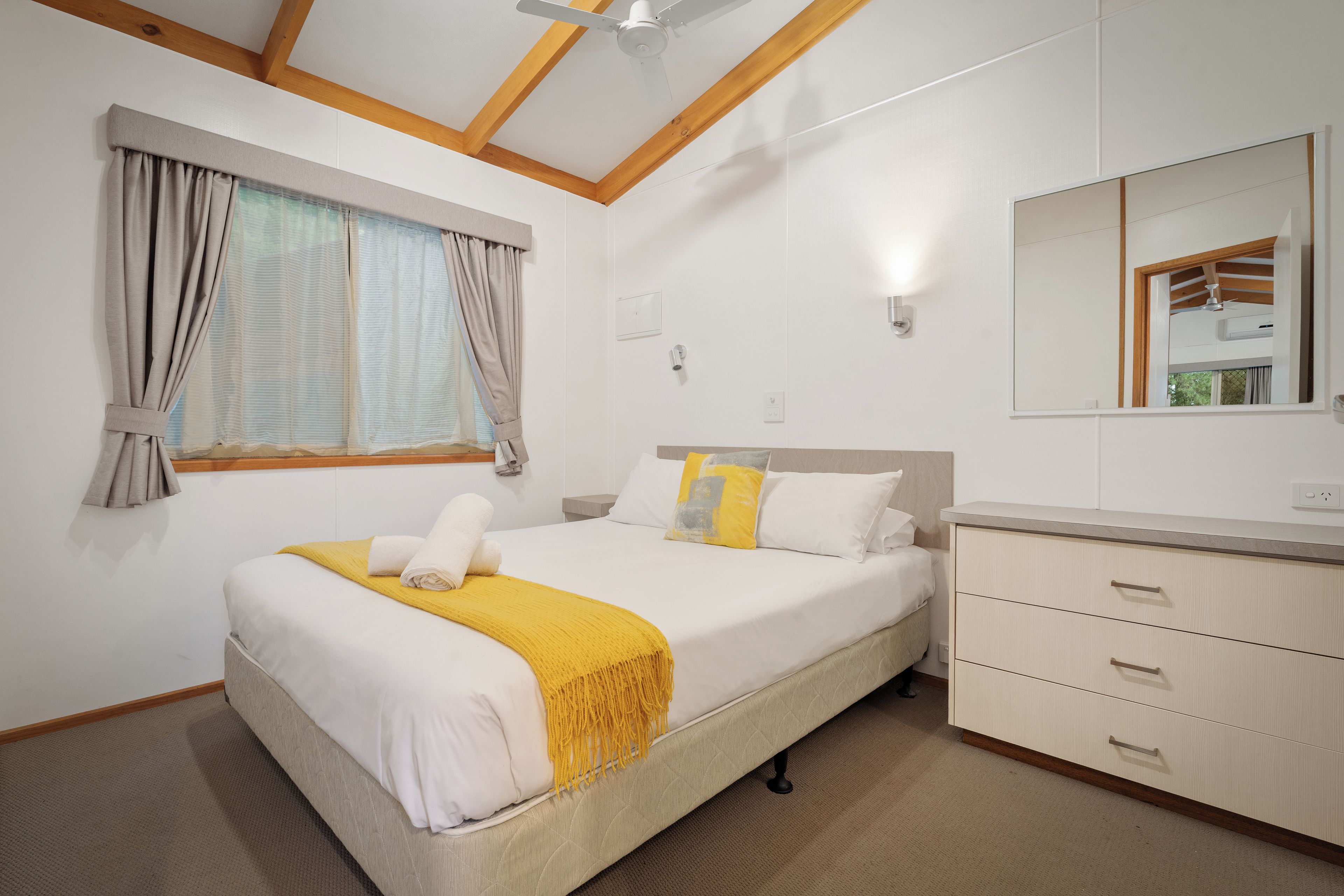 A hotel room with a bed, a bedside table, a dresser, a window with curtains, and a ceiling fan.