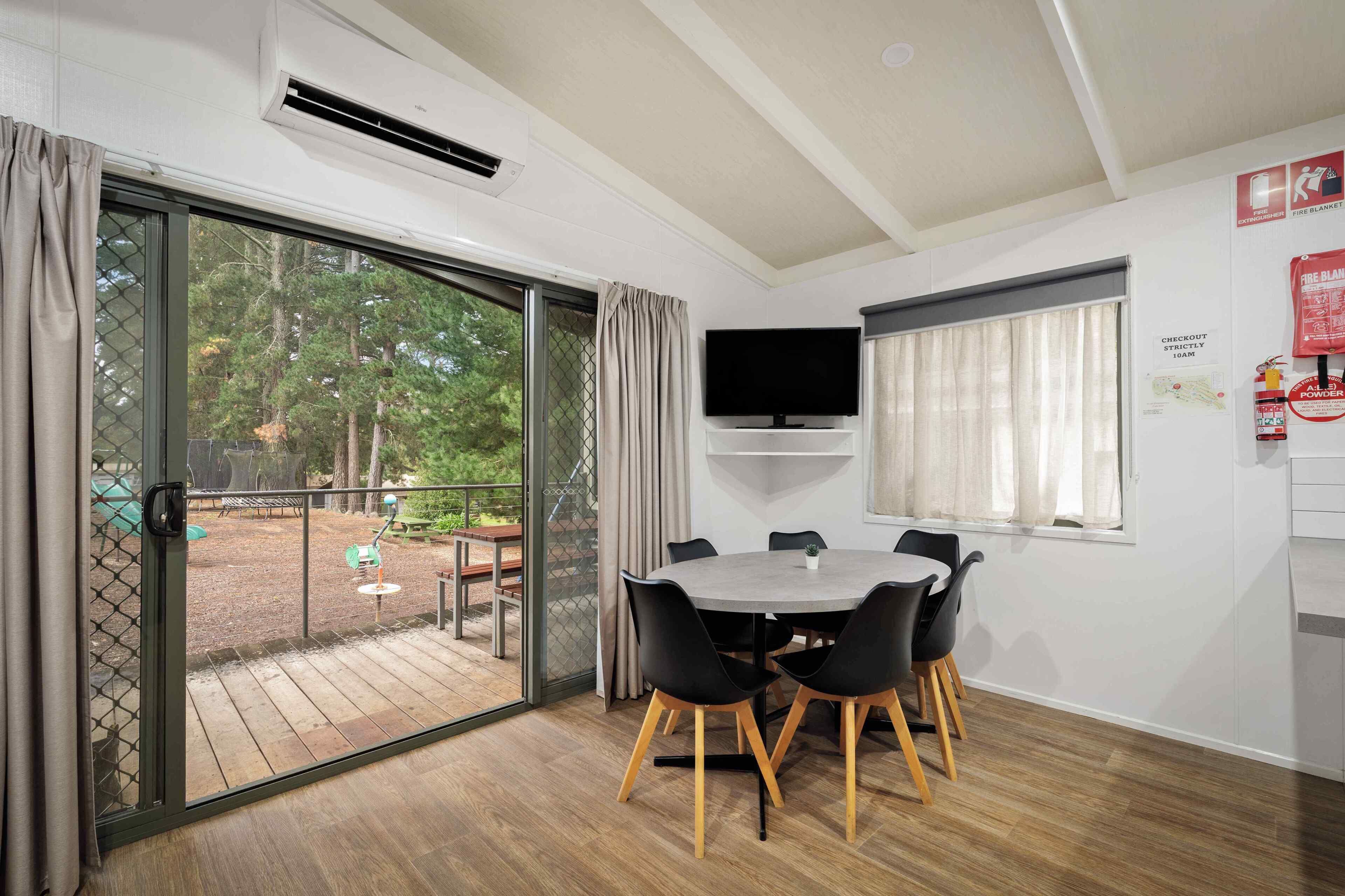 A room with a sliding glass door to an outdoor area, a round table with chairs, a mounted flat-screen TV, and a fire safety notice on the wall.