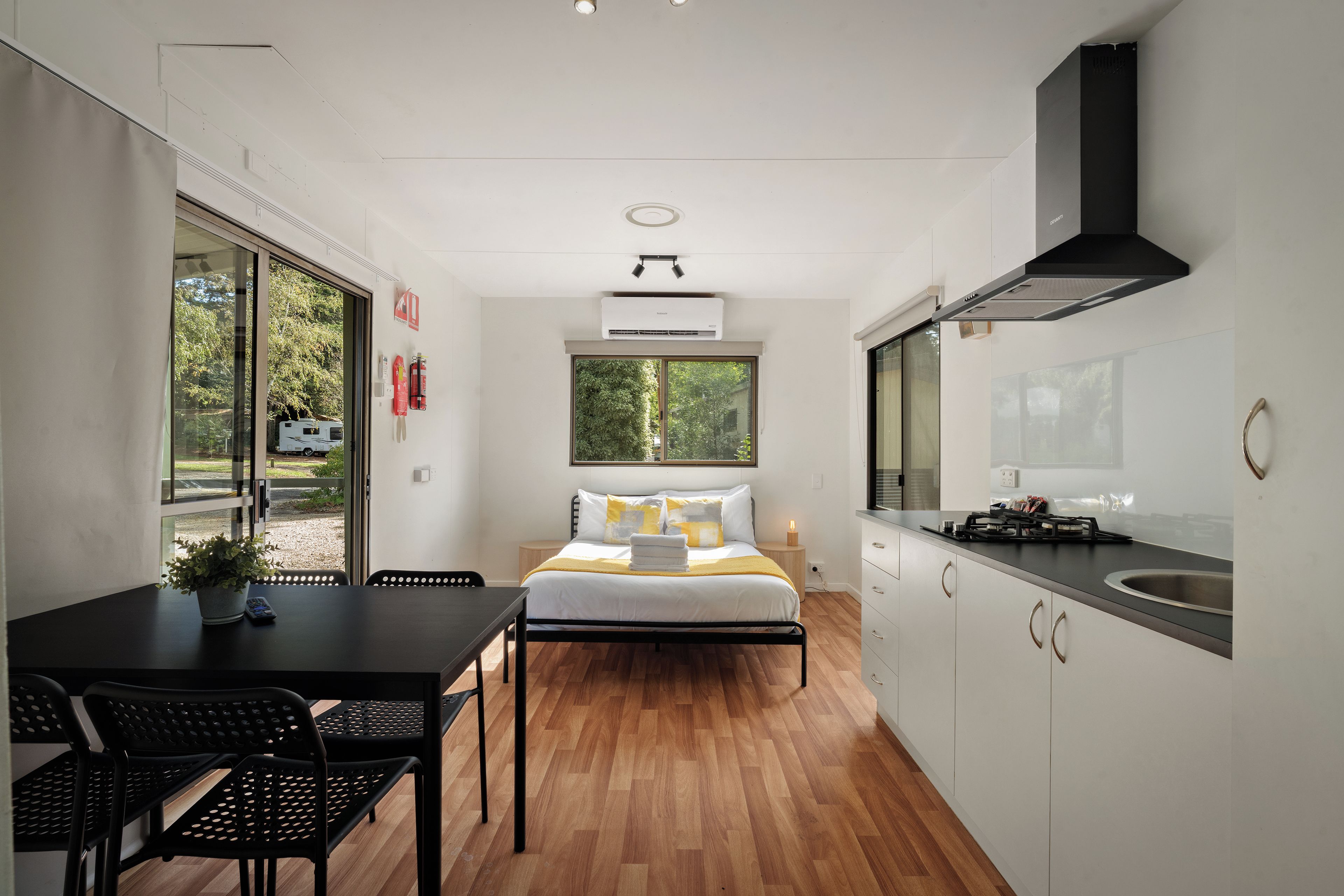 A compact hotel room with a bed, a small table, a chair, a kitchenette, and a window with a view of greenery.