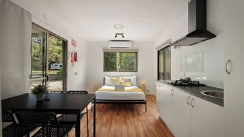 A compact hotel room with a bed, a small table, a chair, a kitchenette, and a window with a view of greenery.
