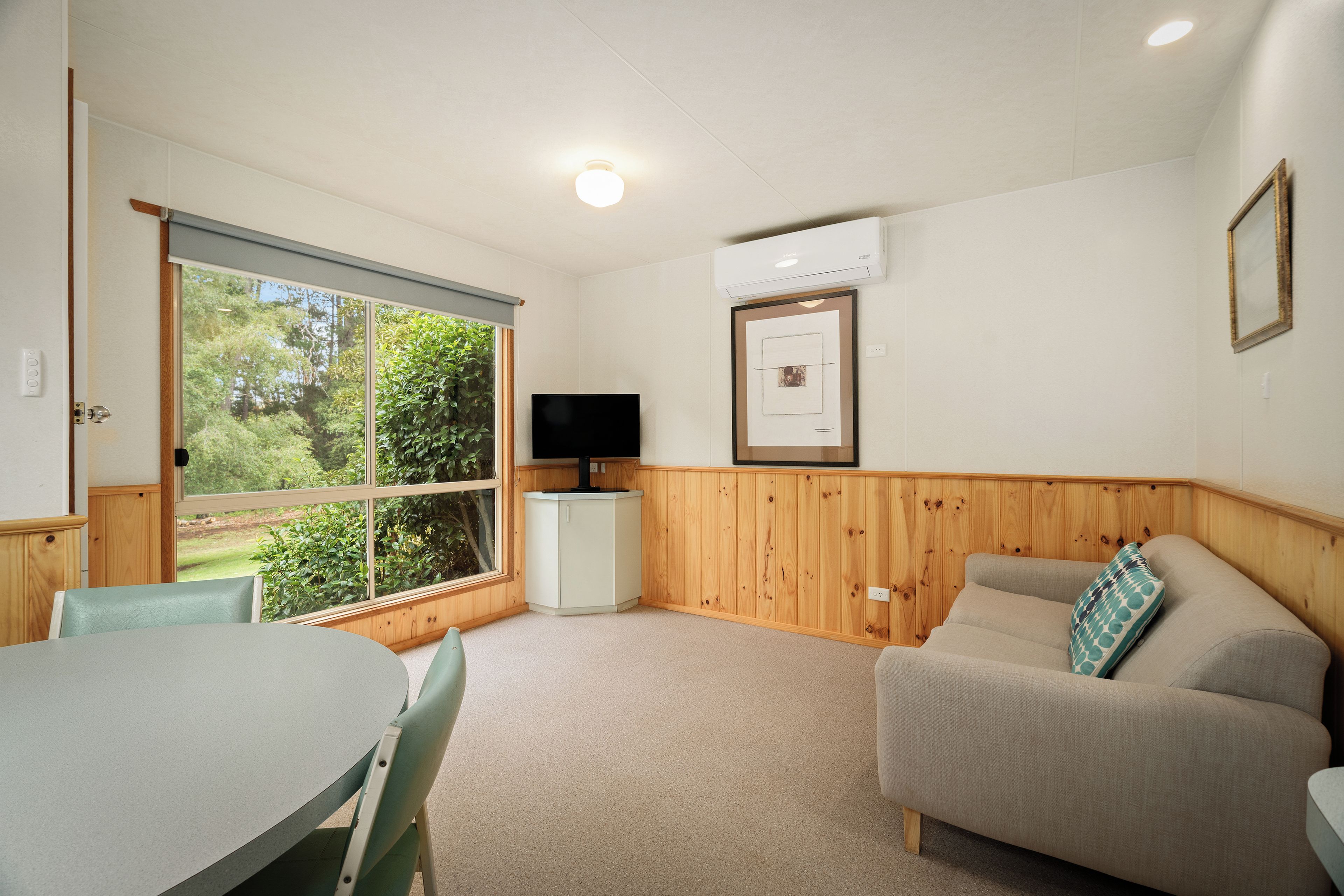 A room with a dining table, a sofa, a television, and a window with a view of greenery.