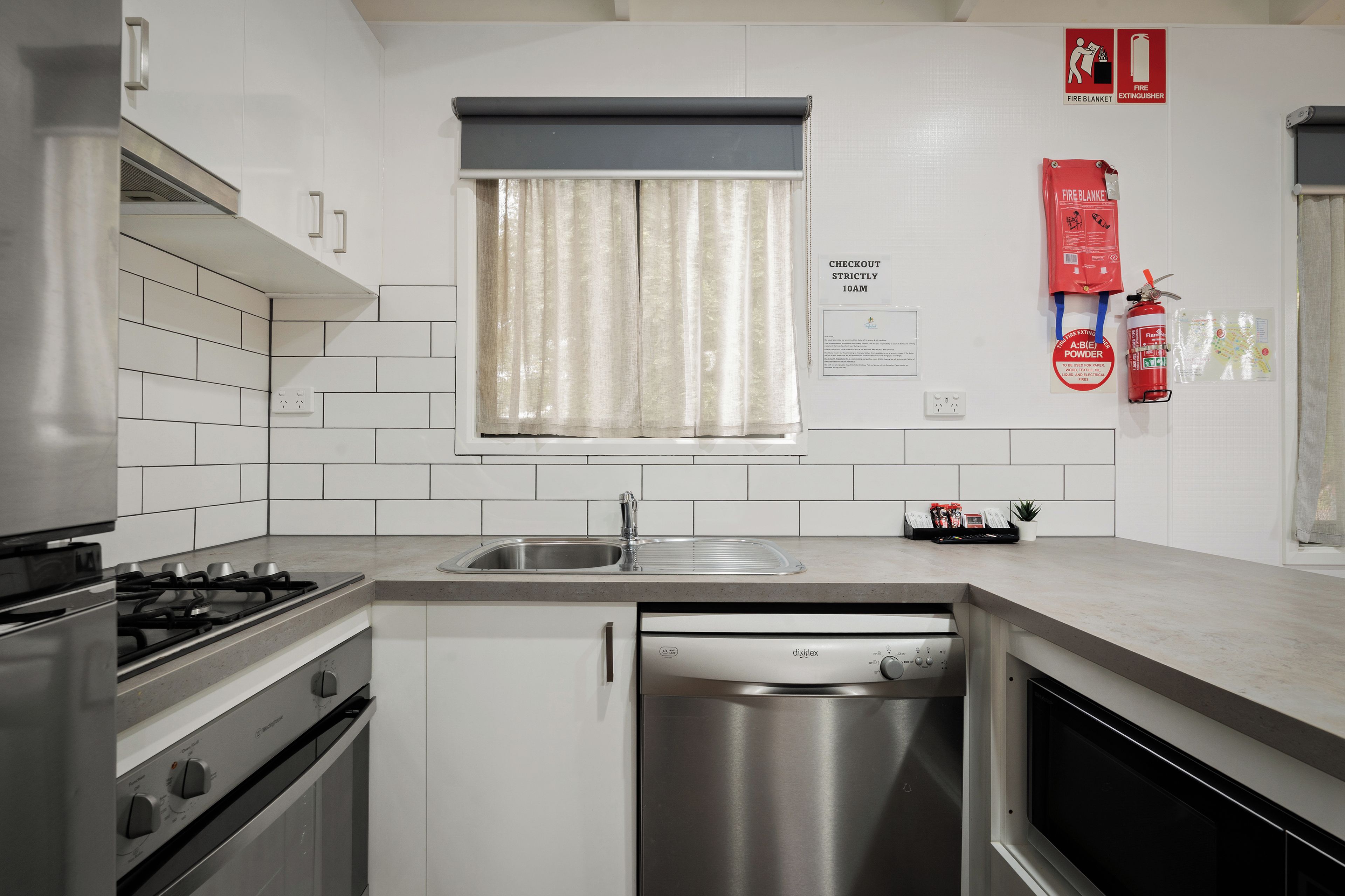 A modern kitchen with white tiles, stainless steel appliances, a window with curtains, and fire safety equipment.
