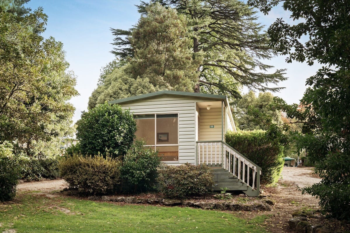 A small, single-story building with a porch, surrounded by dense trees and a grassy area.